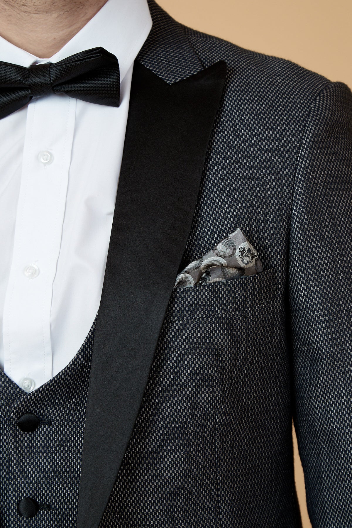 A close-up of a person wearing the marcdarcy SPENCER charcoal grey tux lapel three piece suit, styled with a white dress shirt, black bow tie, and a patterned pocket square in the breast pocket.