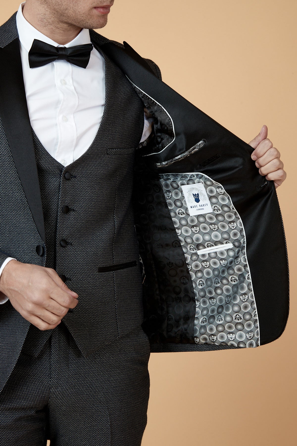 A man models the marcdarcy SPENCER Charcoal Grey Tux Lapel Three Piece Suit, opening his jacket to show the patterned lining and inner white label, styled with a black bow tie against a beige background.