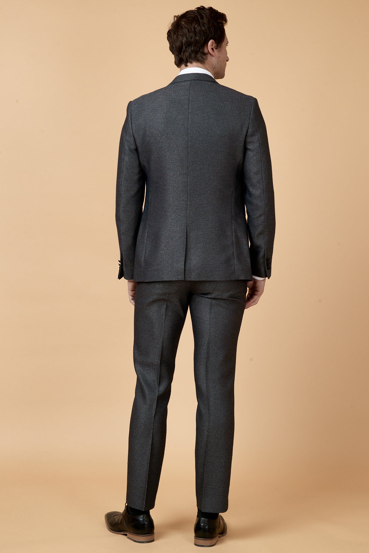 A man with short brown hair wears the marcdarcy SPENCER Charcoal Grey Tux Lapel Three Piece Suit and black shoes, standing with his back to the camera against a plain beige background.