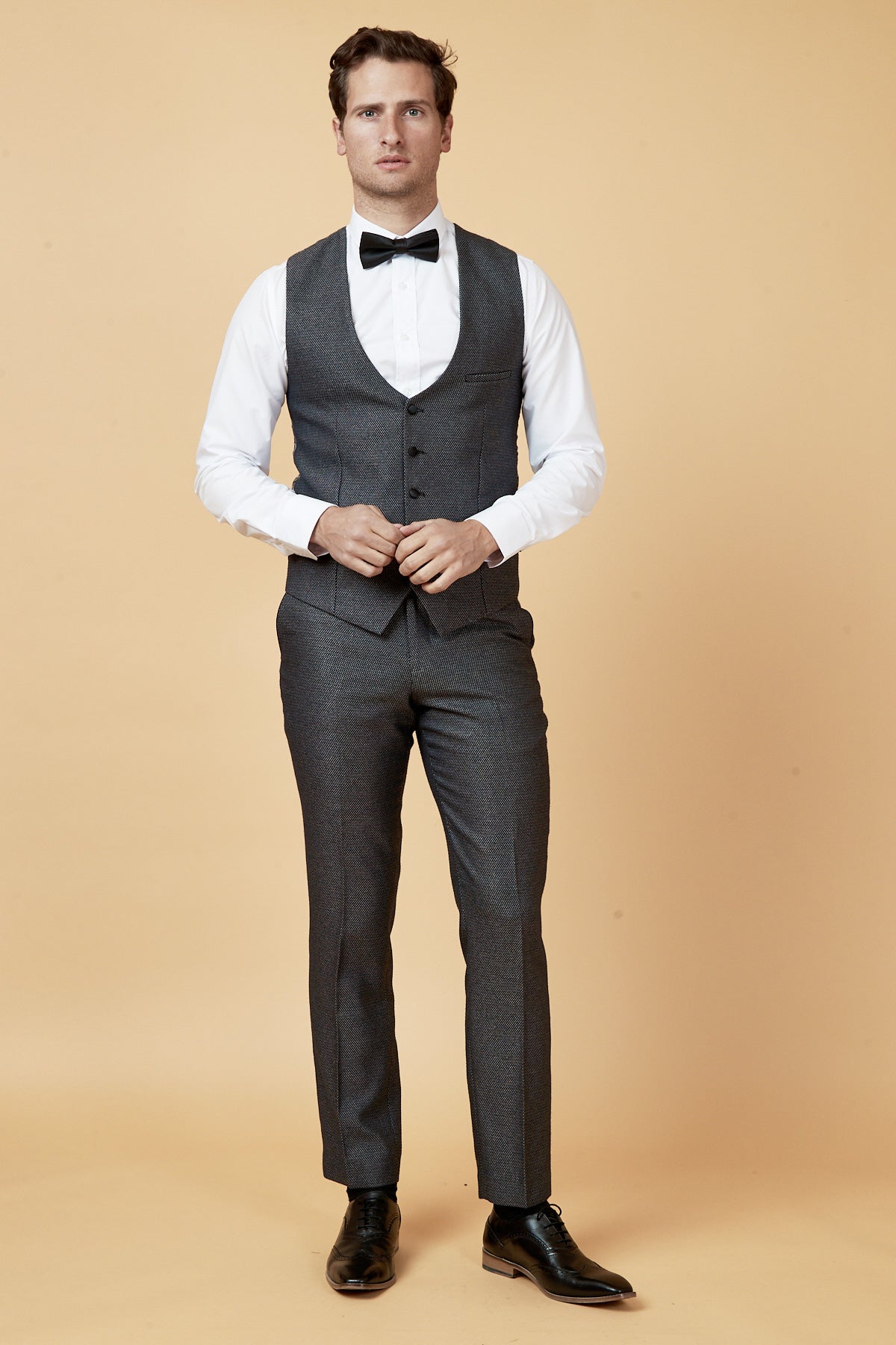 A man stands against a beige background wearing the marcdarcy SPENCER Charcoal Grey Tux Lapel Three Piece Suit with a white dress shirt, black bow tie, and black dress shoes. He faces forward, hands clasped in front of his waist.
