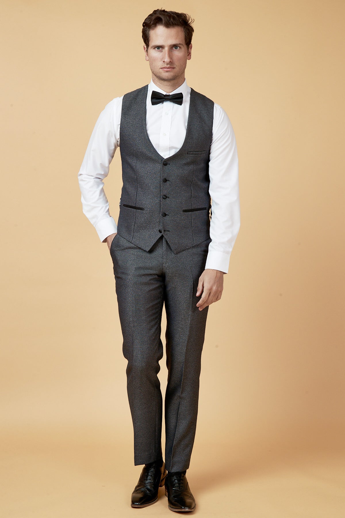 A man stands against a beige background wearing the marcdarcy SPENCER Charcoal Grey Tux Lapel Three Piece Suit with a white dress shirt, black bow tie, and black dress shoes, hands in pockets and looking straight ahead.