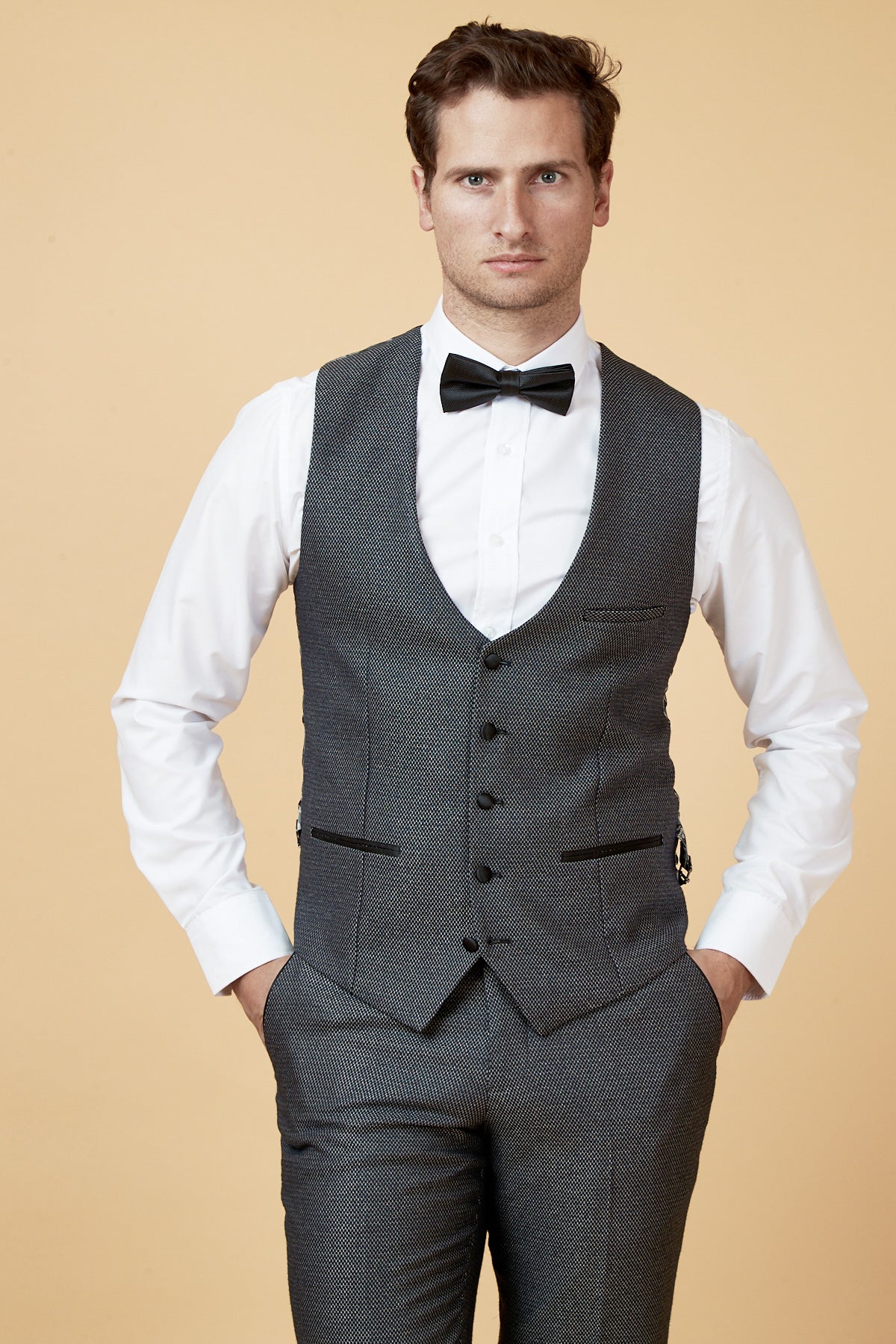 A man stands against a beige background wearing the marcdarcy SPENCER Charcoal Grey Single Breasted Waistcoat, paired with a matching suit, white dress shirt, and black bow tie. His hands are in his pockets as he looks at the camera.