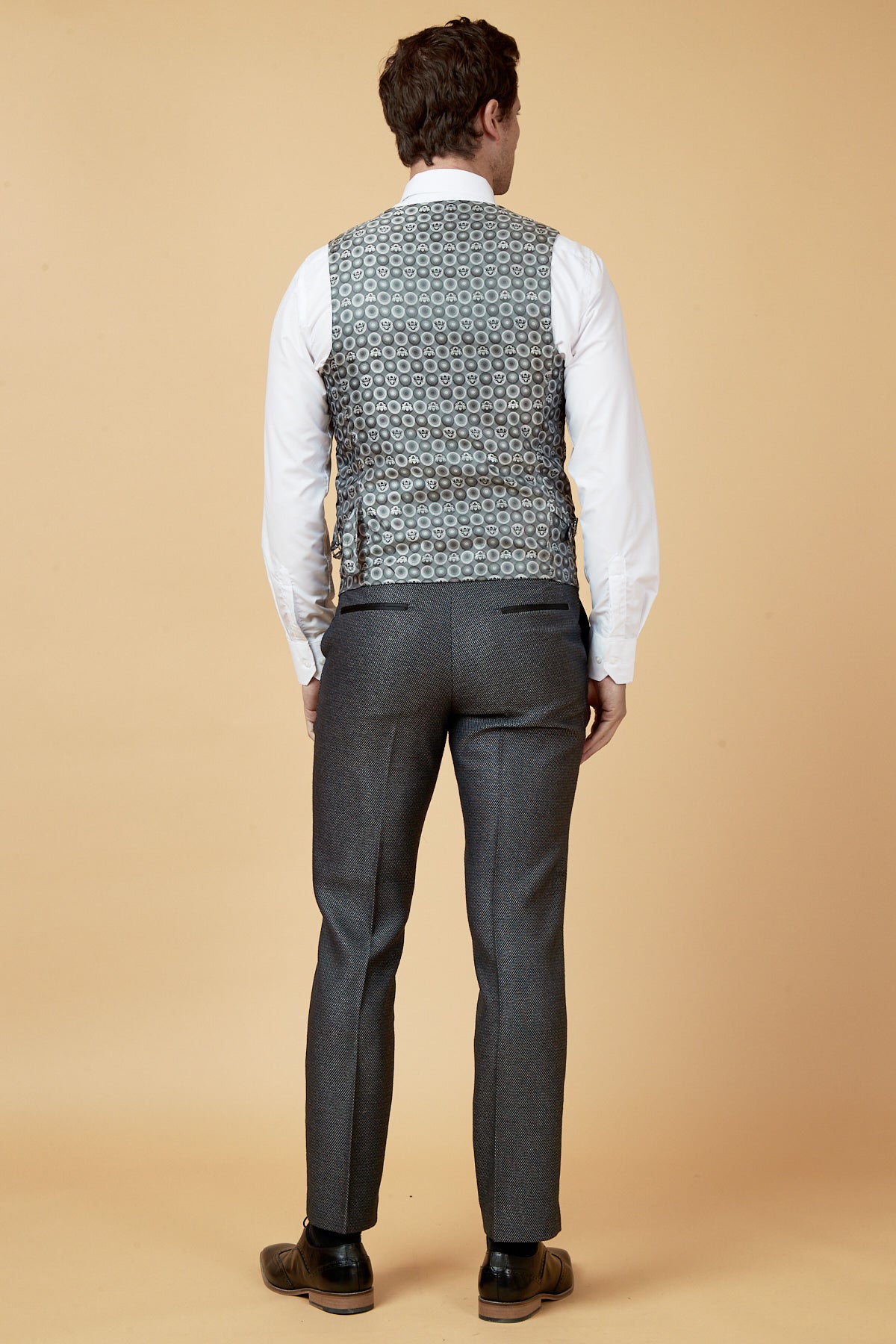 A man faces away against a beige background, wearing the marcdarcy SPENCER Charcoal Grey Tux Lapel Three Piece Suit with slim fit pants, a white shirt, black shoes, and a geometric patterned vest.