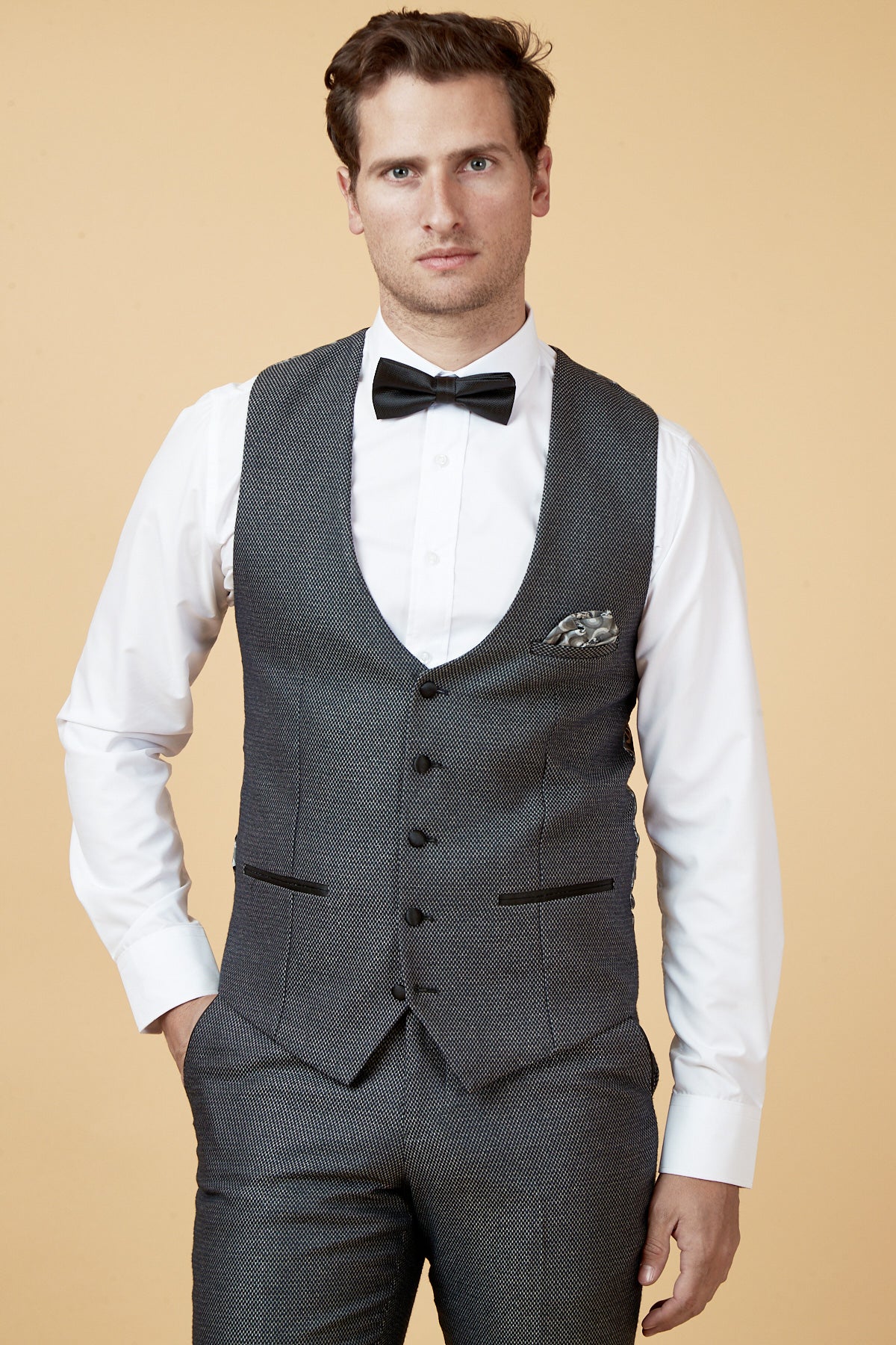 A man in a marcdarcy SPENCER Charcoal Grey Tux Lapel Three Piece Suit stands with his hand in his pocket against a peach background, wearing a white shirt and black bow tie.
