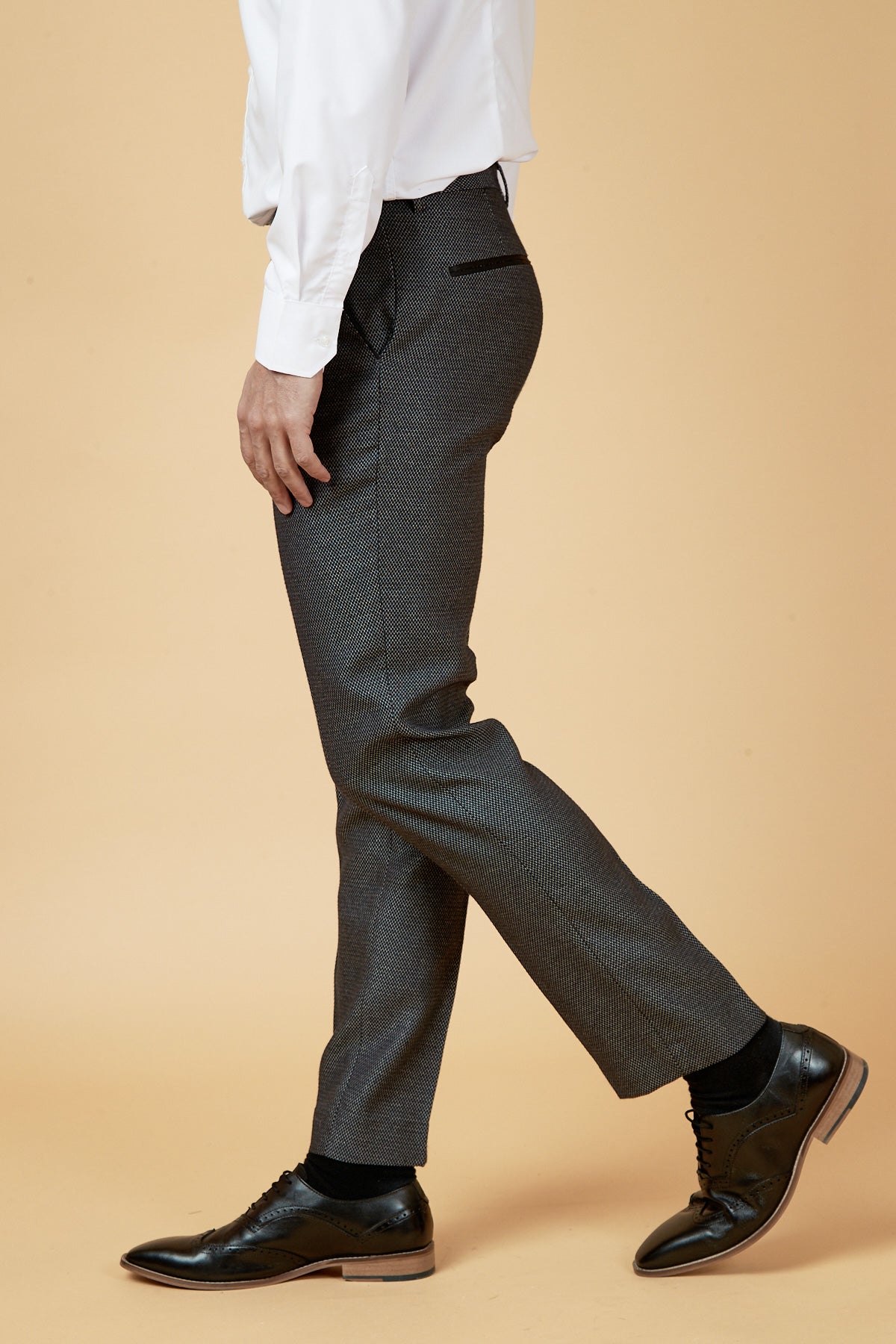A person is shown standing sideways in a beige setting, wearing the marcdarcy SPENCER - Charcoal Grey Tux Lapel Three Piece Suit. The outfit includes a slim fit white dress shirt, charcoal suit pants, black socks, and black dress shoes; head not visible.
