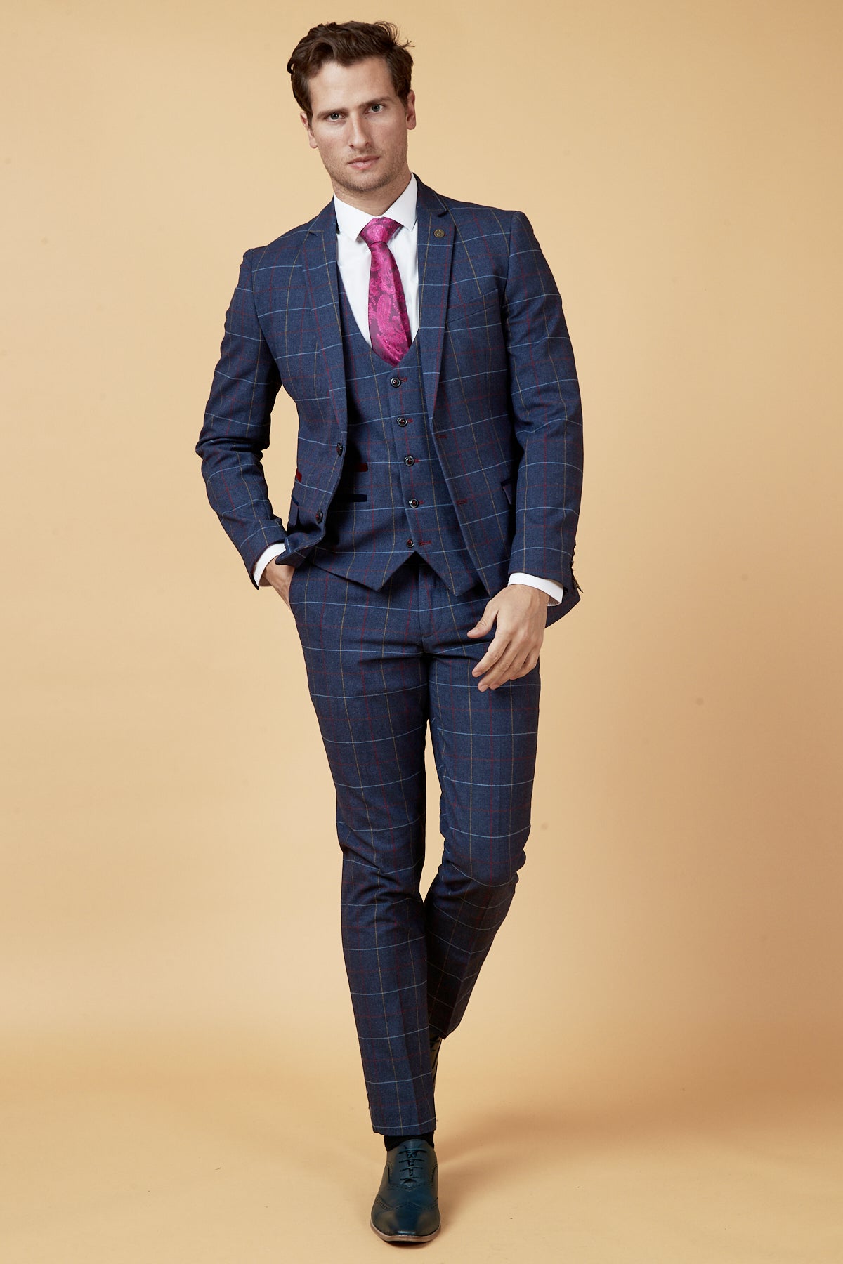 A man wearing the marcdarcy DRAKE navy check three piece suit, a white dress shirt, and a bright pink tie stands against a beige background with one leg crossed in front of the other and one hand in his pocket.