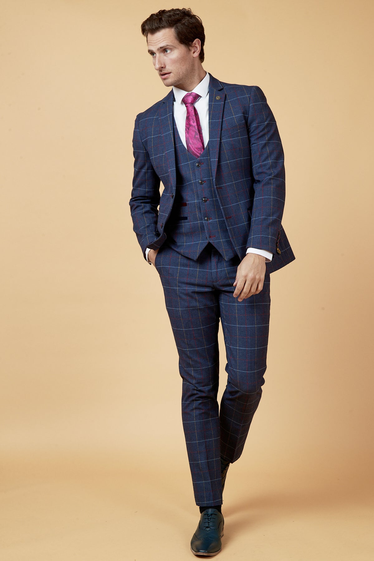 A man wearing the marcdarcy DRAKE Navy Check Three Piece Suit with a white shirt and a pink patterned tie stands with his hand in his pocket, ideal for any formal event.