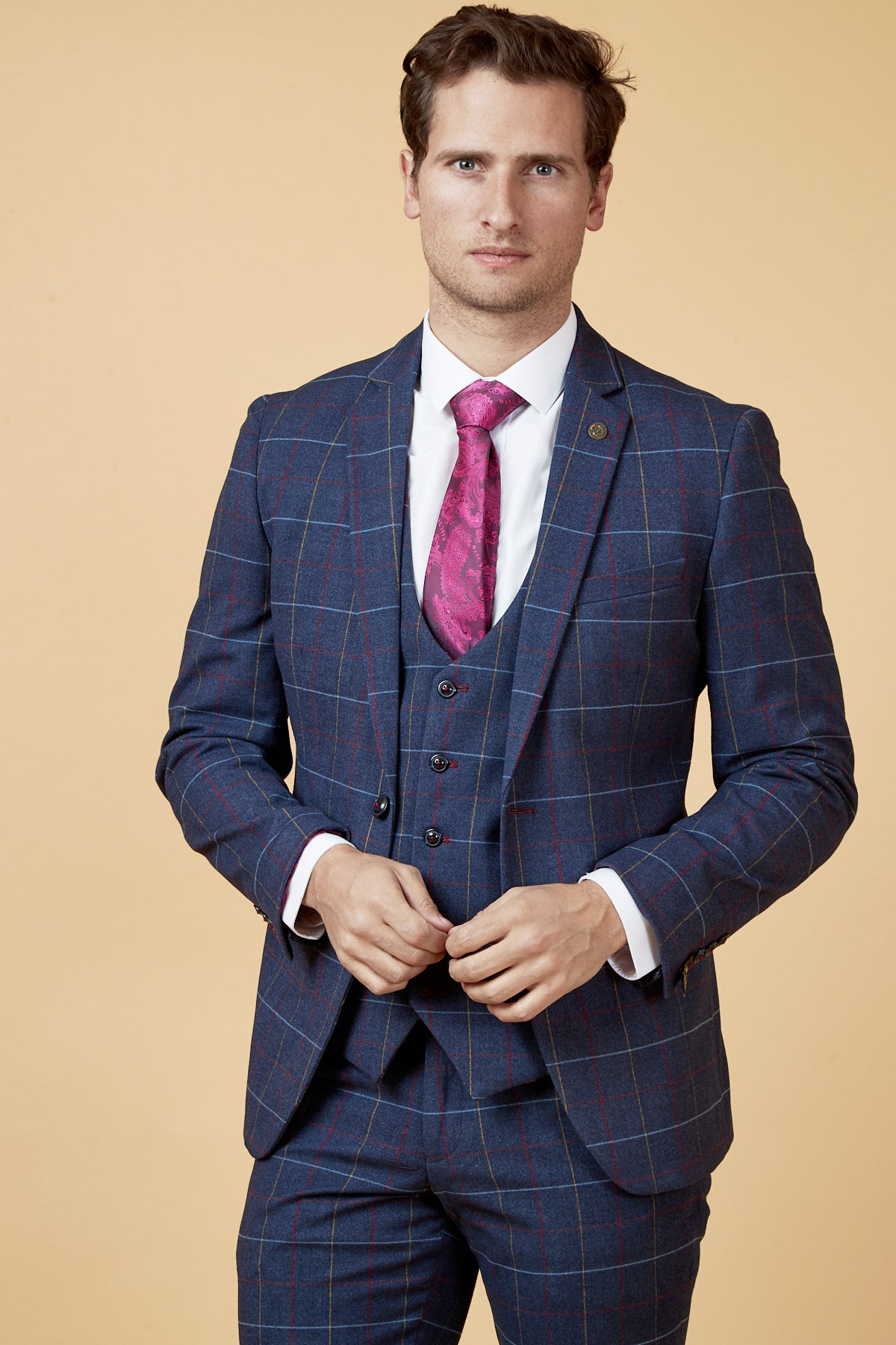 A man in a marcdarcy DRAKE Navy Check Three Piece Suit, styled with a white shirt and pink patterned tie, stands against a beige background, looking at the camera with a neutral expression.