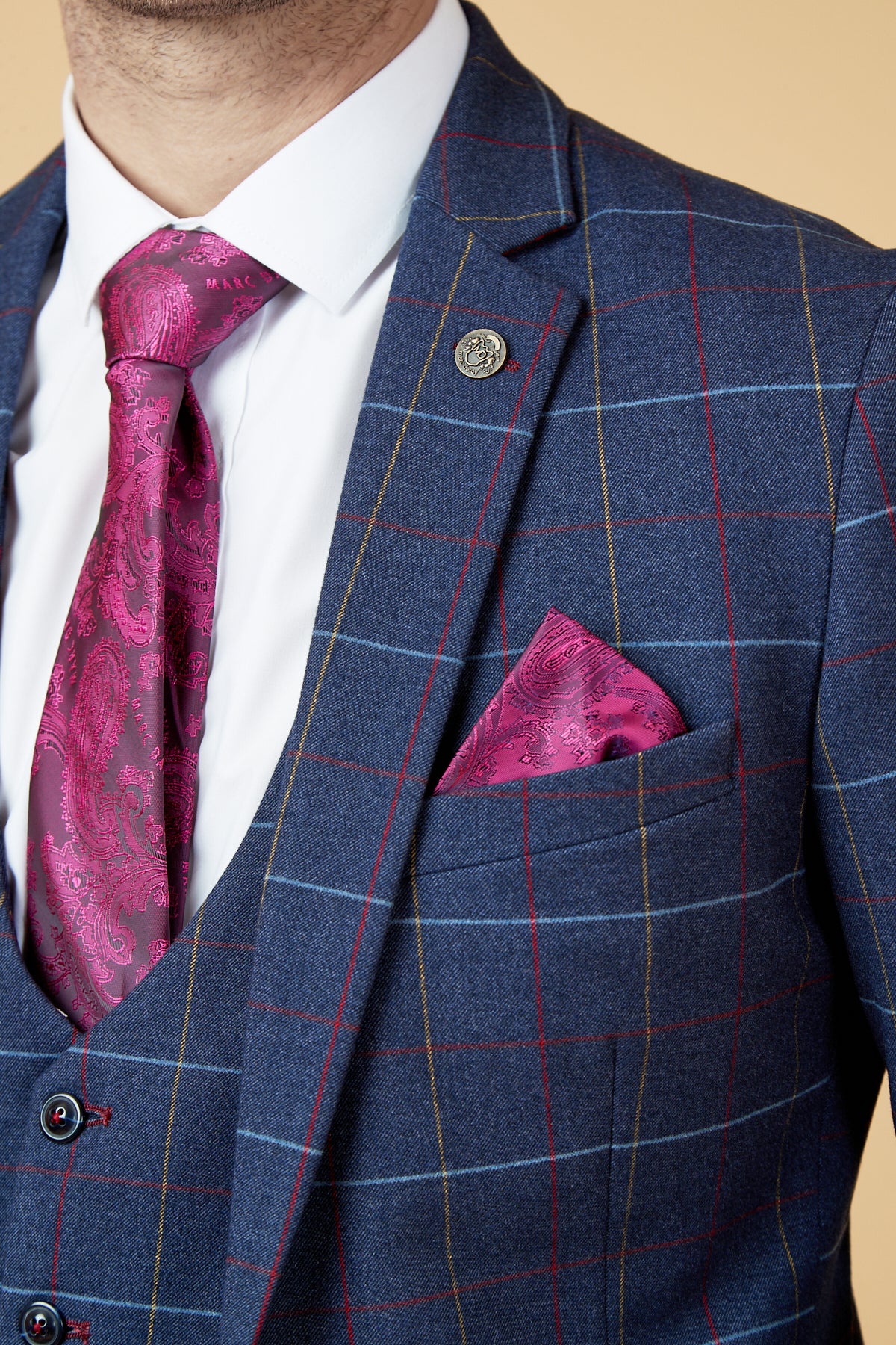A man wears the marcdarcy DRAKE navy check three piece suit with red and yellow lines, a white shirt, magenta paisley tie and matching pocket square, plus a lapel pin—all set against a beige background.