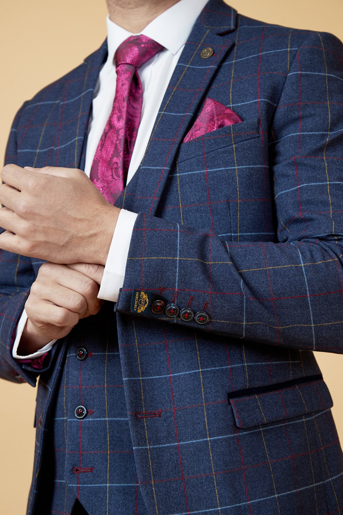 A man adjusts his white shirt cuff while wearing the DRAKE Navy Check Three Piece Suit by marcdarcy, paired with a pink patterned tie and matching pocket square, set against a beige background.