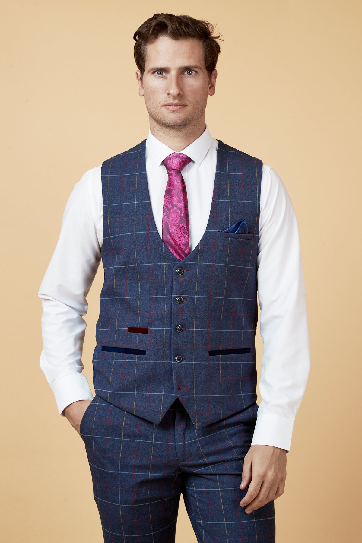 A man wearing the marcdarcy DRAKE - Navy Check Waistcoat and matching trousers, paired with a white shirt and pink patterned tie, stands with one hand in his pocket against a beige background.