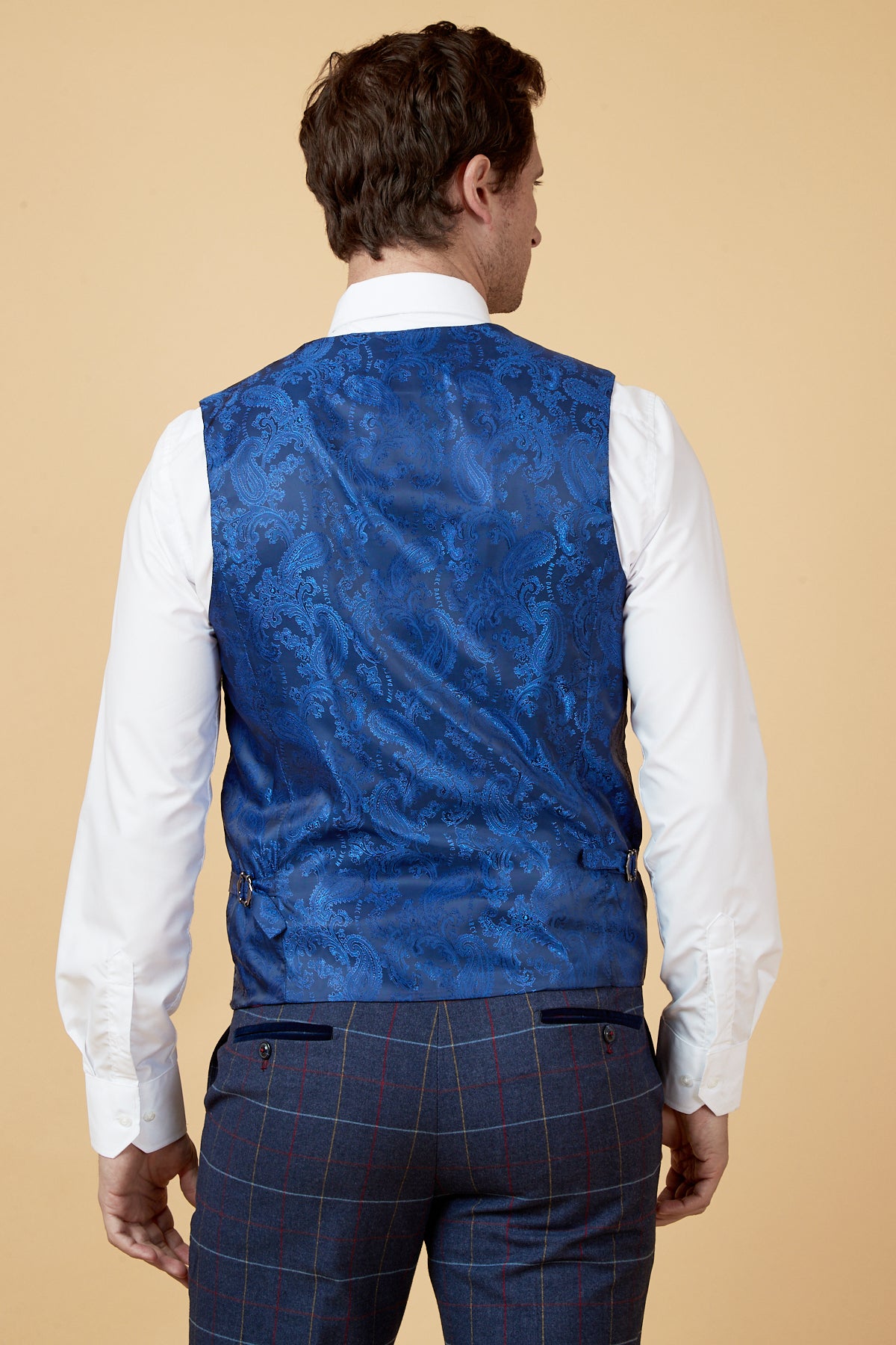 A man facing away wears a white dress shirt, the marcdarcy DRAKE - Navy Check Waistcoat, and plaid trousers with a beige background.