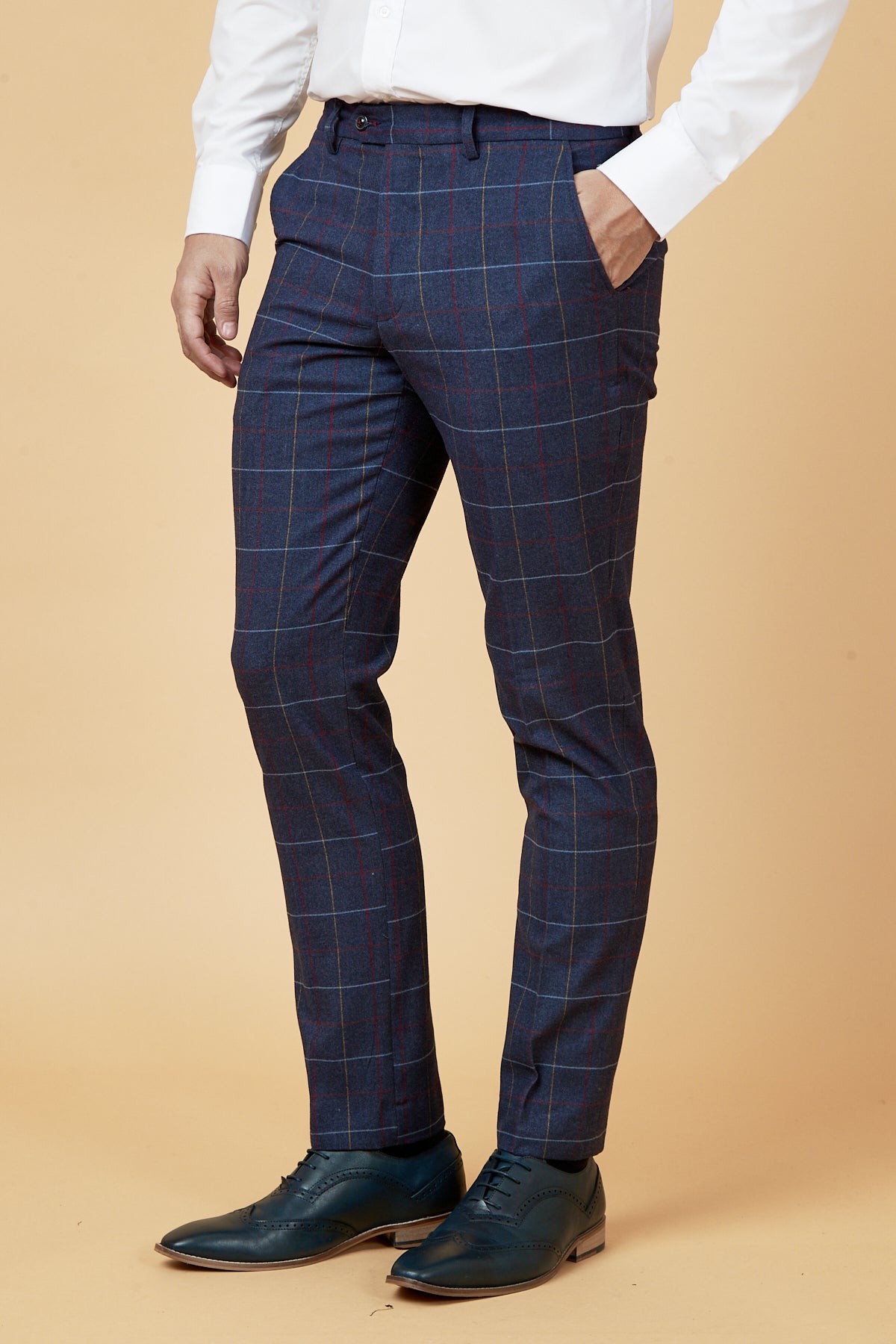 A man stands against a beige background with one hand in his pocket, wearing marcdarcy's DRAKE - Navy Check Trousers, a white dress shirt, and dark green dress shoes.