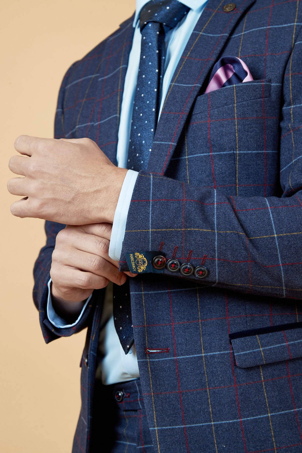 A man in the marcdarcy DRAKE Navy Check Blazer adjusts his cuff. He pairs it with a light blue shirt, navy polka dot tie, and a pink pocket square against a plain beige background.
