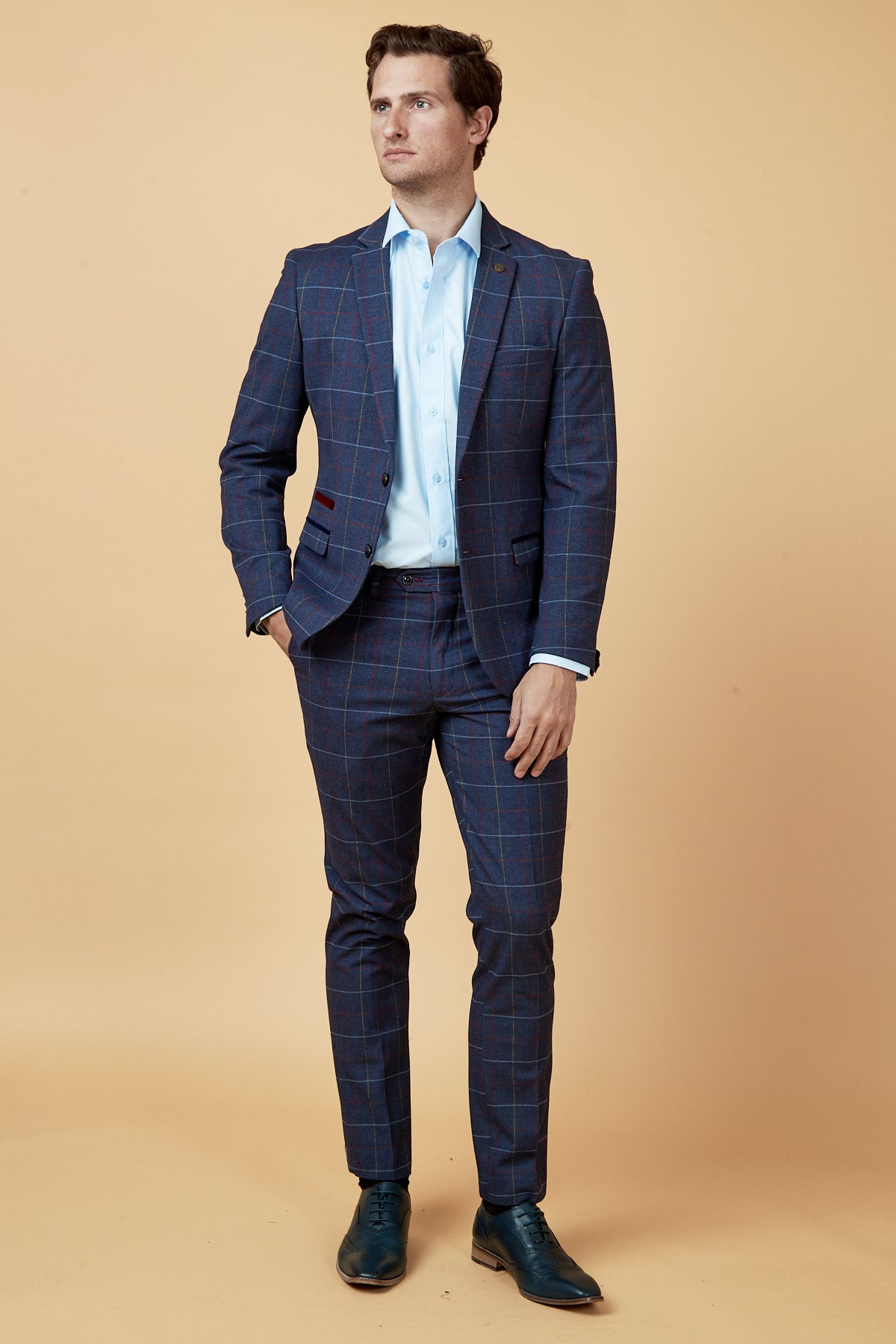 A man with short brown hair stands against a beige background, wearing the marcdarcy DRAKE - Navy Check Blazer, a light blue dress shirt, and dark blue dress shoes. He looks slightly to the side.