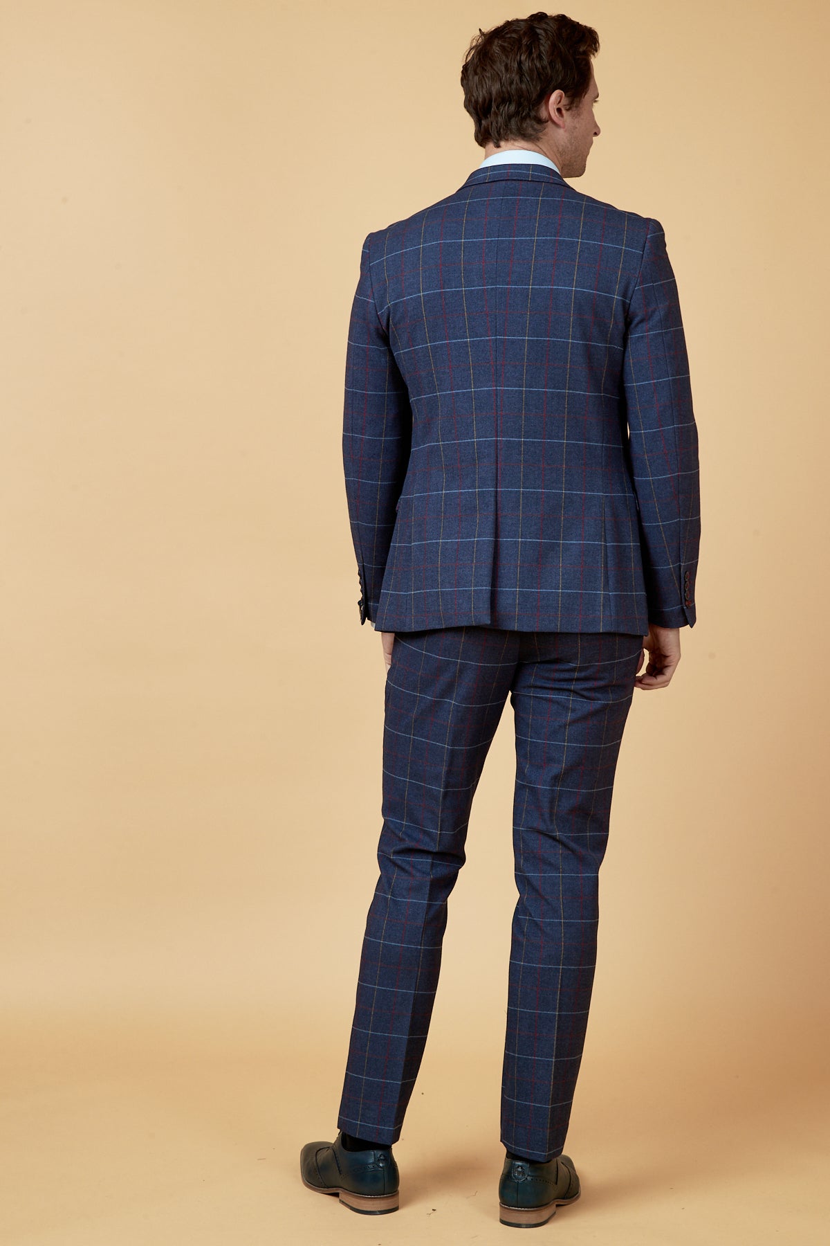 A man faces away, wearing the marcdarcy DRAKE - Navy Check Blazer with red and white lines, a white shirt, and black dress shoes against a plain beige background.