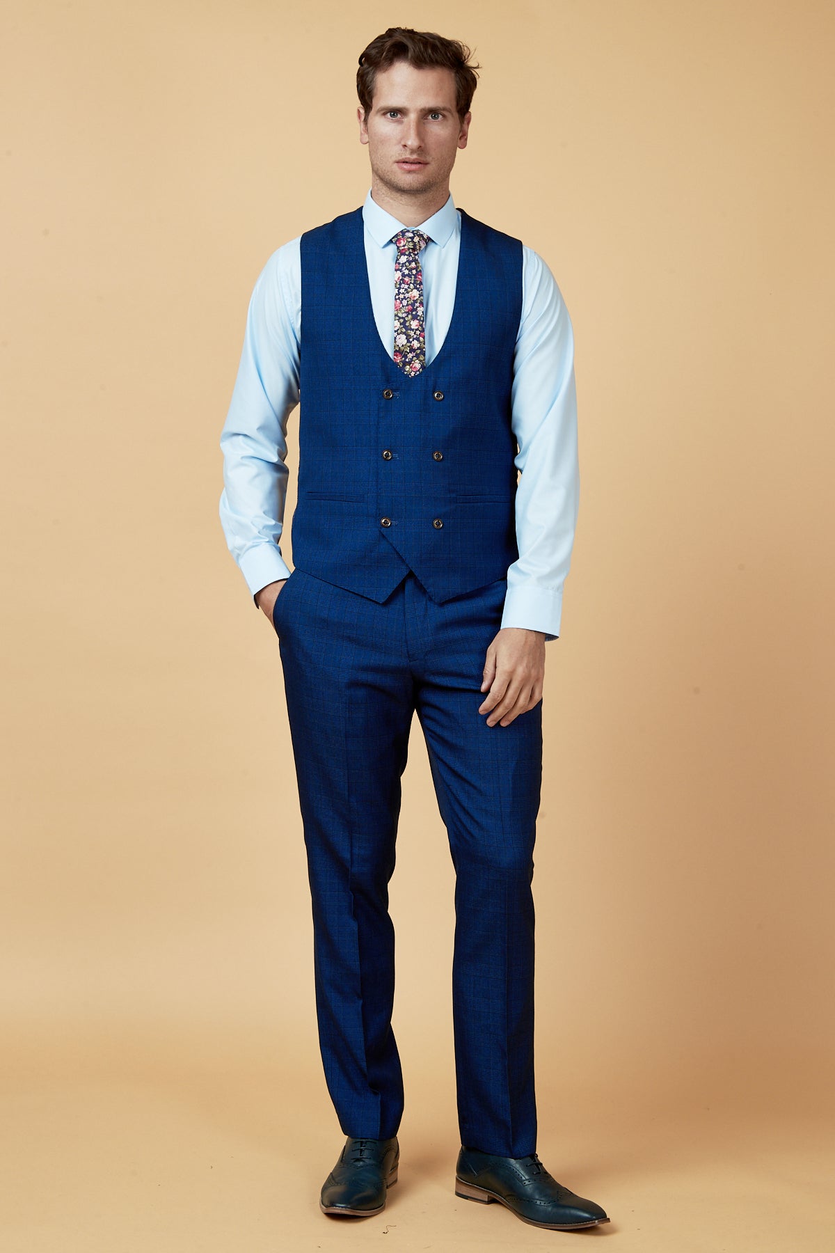 A man stands against a beige background, wearing the marcdarcy LEE GEORGE Royal Blue Double Breasted Waistcoat, a light blue shirt, floral tie, and dark blue dress shoes. He looks straight ahead with a neutral expression.