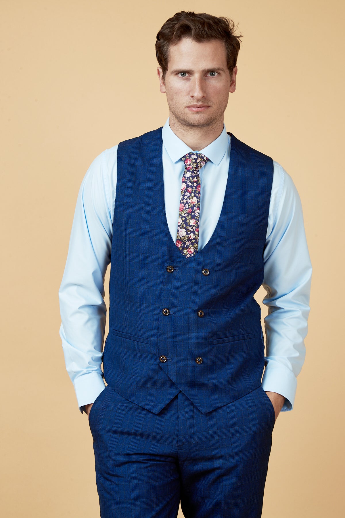 A man models the marcdarcy LEE GEORGE Royal Blue Double Breasted Waistcoat, paired with matching trousers, a light blue shirt, and a floral tie, presenting elegant formalwear against a beige backdrop.