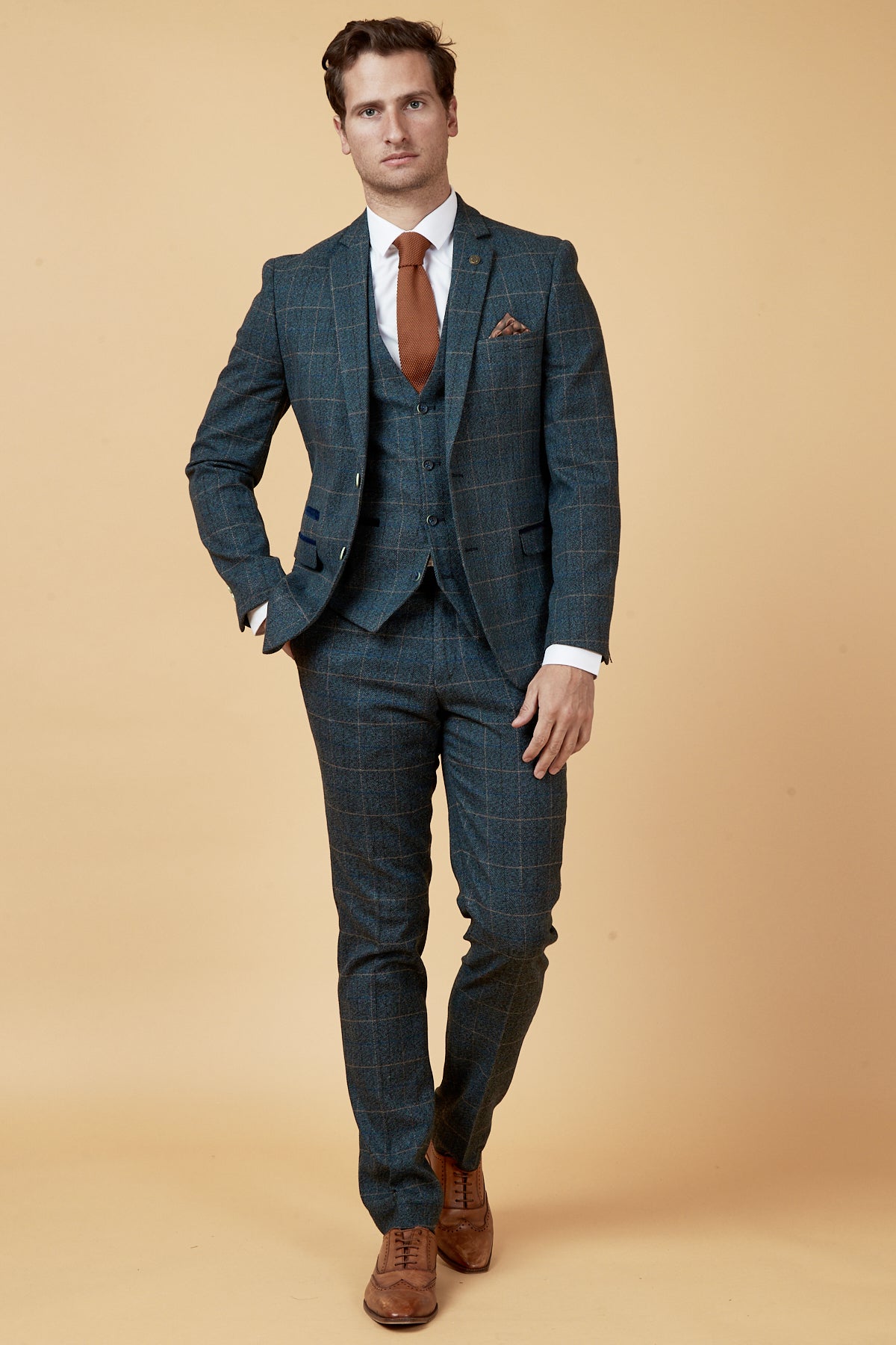 A man in a Marc Darcy SCOTT blue check three piece suit, white shirt, brown tie, and brown shoes stands against a beige background with one hand in his pocket, channeling classic Peaky Blinders style.