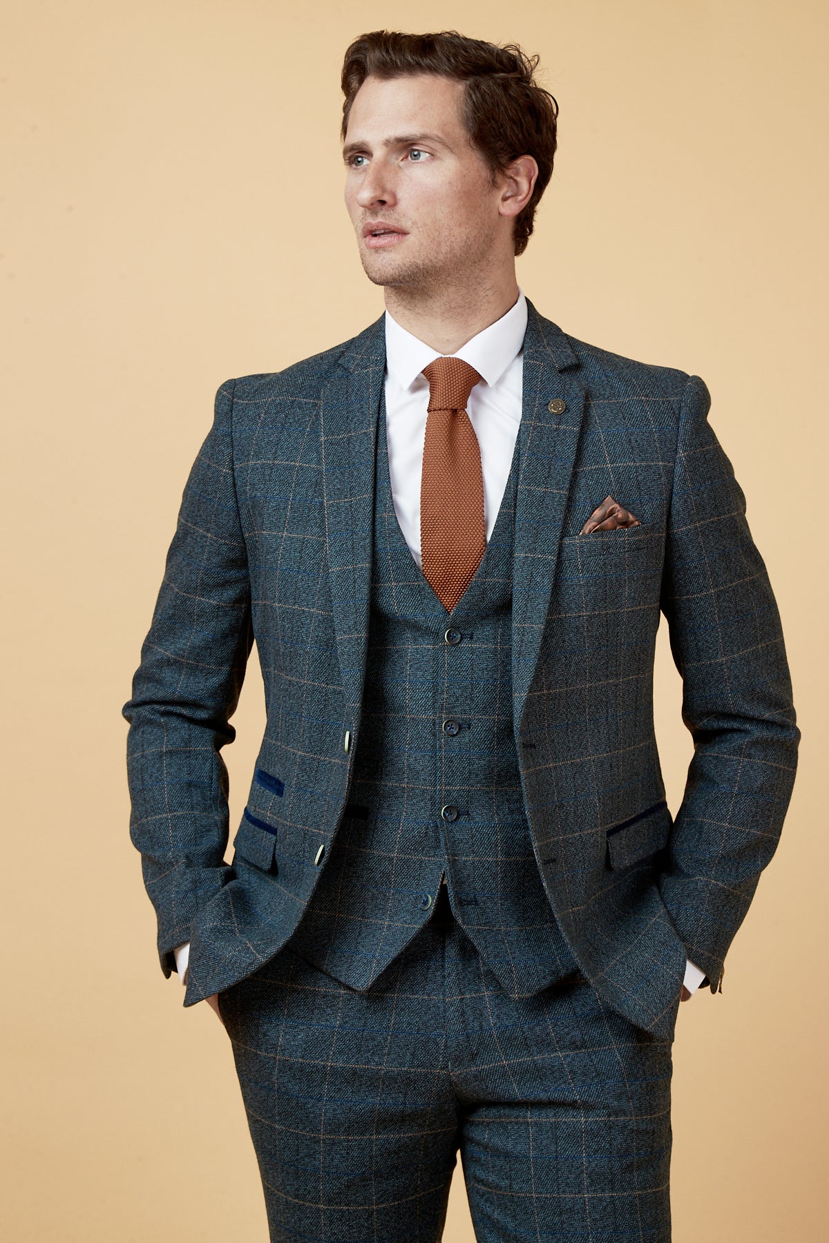 A man wears the Marc Darcy SCOTT Blue Check Three Piece Suit with a white shirt and burnt orange tie, standing with hands in pockets against a beige background.
