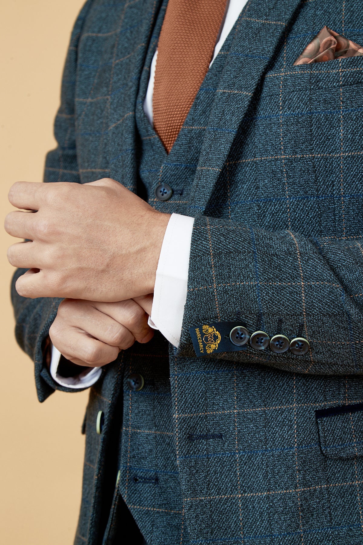 A person in the marcdarcy SCOTT - Blue Tweed Check Blazer with a white shirt and brown tie adjusts their shirt cuff, showing only their upper body and hands against a beige background.