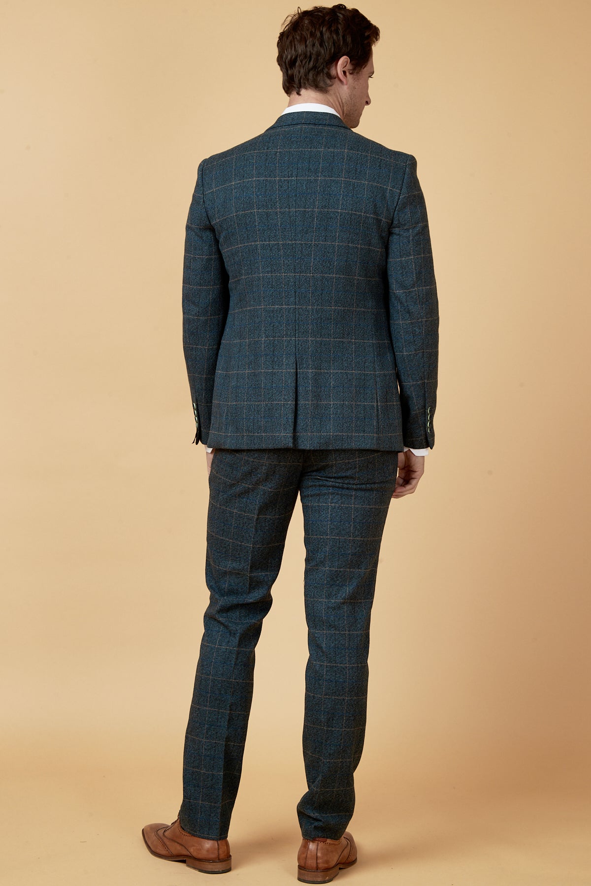 A man with short brown hair stands with his back turned, wearing the Marc Darcy SCOTT - Blue Check Three Piece Suit and brown shoes against a beige background, evoking a vintage-inspired style.