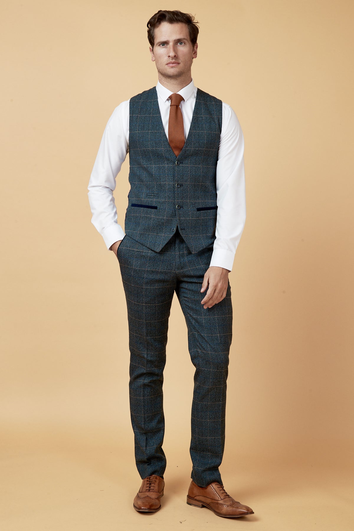 A man stands against a beige background wearing the Marc Darcy SCOTT Blue Check Three Piece Suit, paired with a white shirt, rust tie, and brown shoes, channeling classic Peaky Blinders style as he faces forward with his hands at his sides.