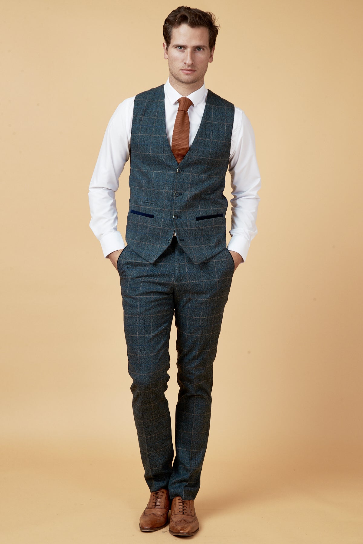 A man stands against a beige background wearing the marcdarcy LEN SCOTT Blue Tweed Check Waistcoat and matching pants, paired with a white shirt, brown tie, and brown dress shoes, hands in pockets, highlighting a vintage-inspired style.