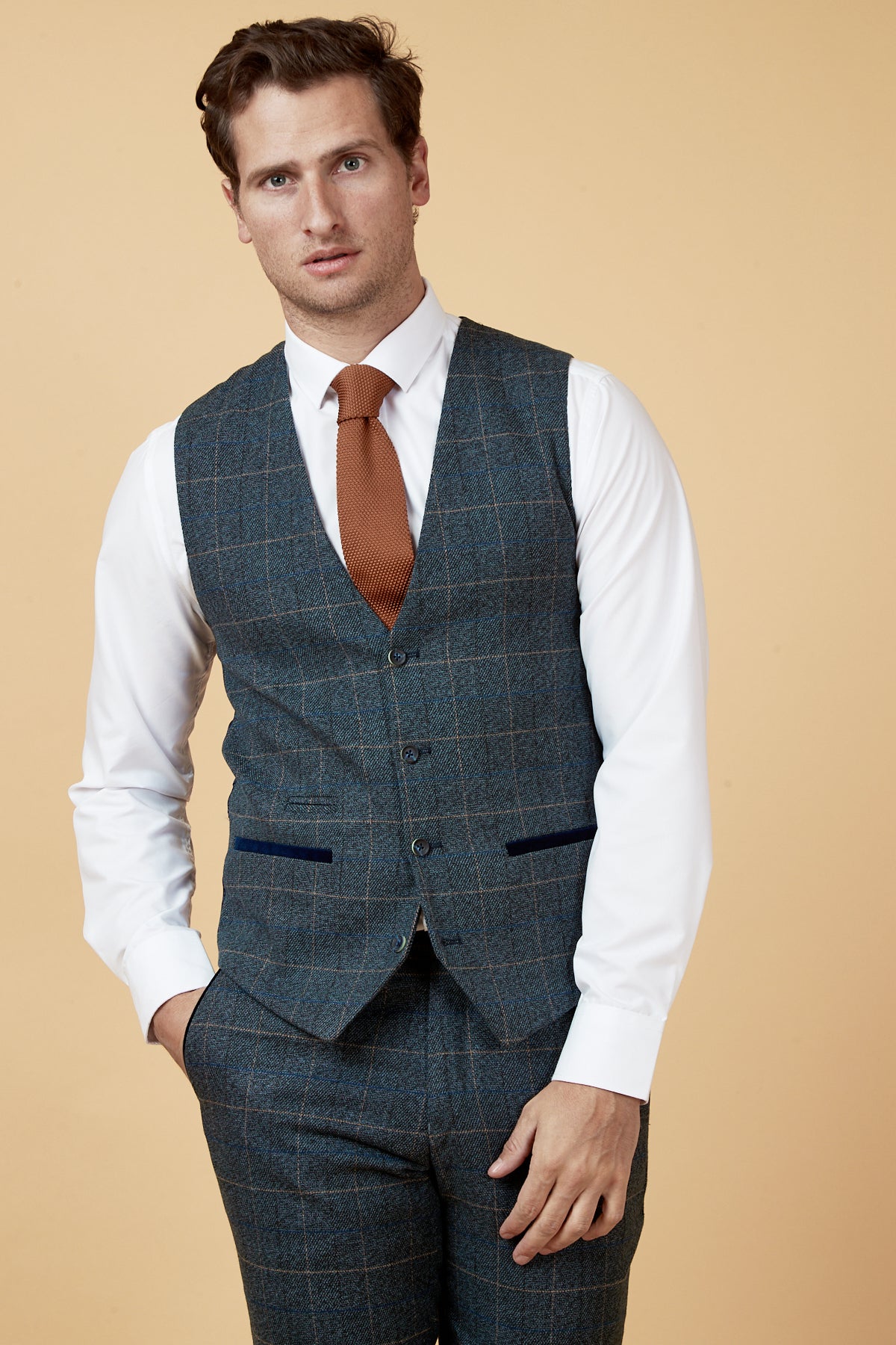 A man wears the marcdarcy LEN SCOTT Blue Tweed Check Waistcoat over a white shirt and orange tie, standing against a cream background with one hand in his pocket.