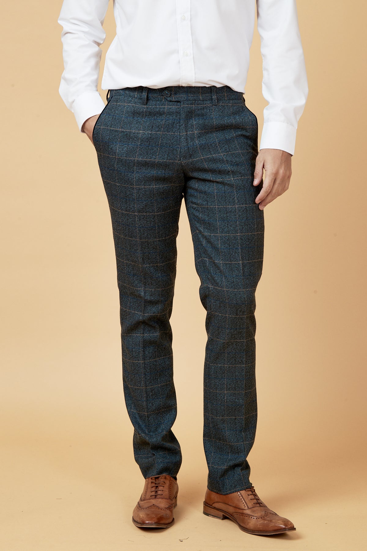 A man in a white dress shirt, marcdarcy SCOTT Blue Check Tweed Trousers, and brown leather dress shoes stands with one hand in his pocket against a beige background.
