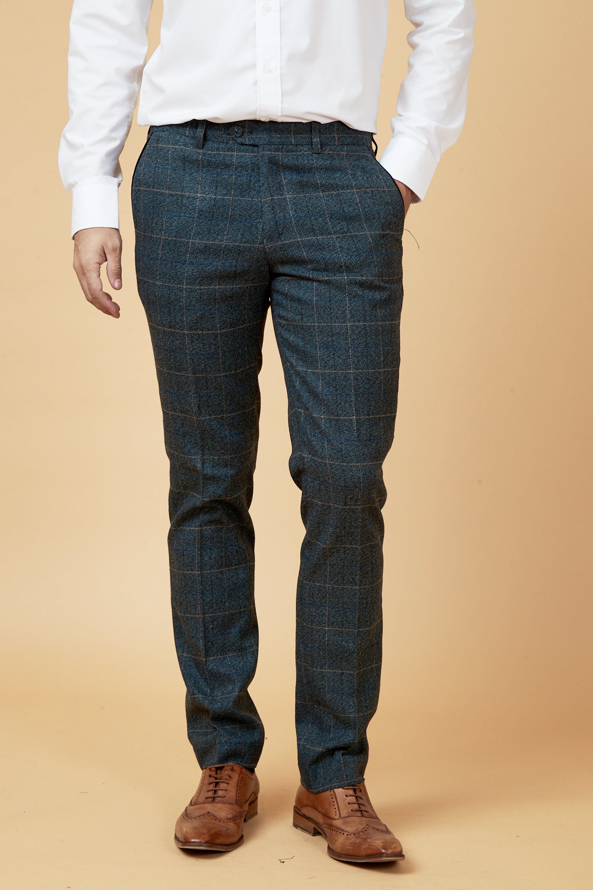 A man wearing a white dress shirt and marcdarcy’s SCOTT - Blue Check Tweed Trousers stands with one hand in his pocket, paired with brown leather dress shoes against a beige background.