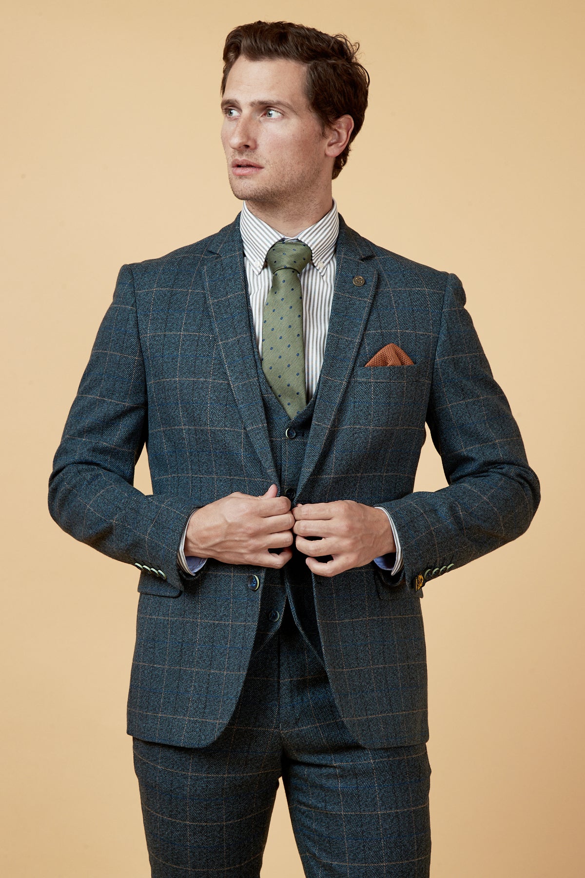 A man wears the marcdarcy SCOTT Blue Tweed Check Blazer with a green polka dot tie, striped shirt, and orange pocket square, standing against a beige background and looking to the side as he buttons his jacket.
