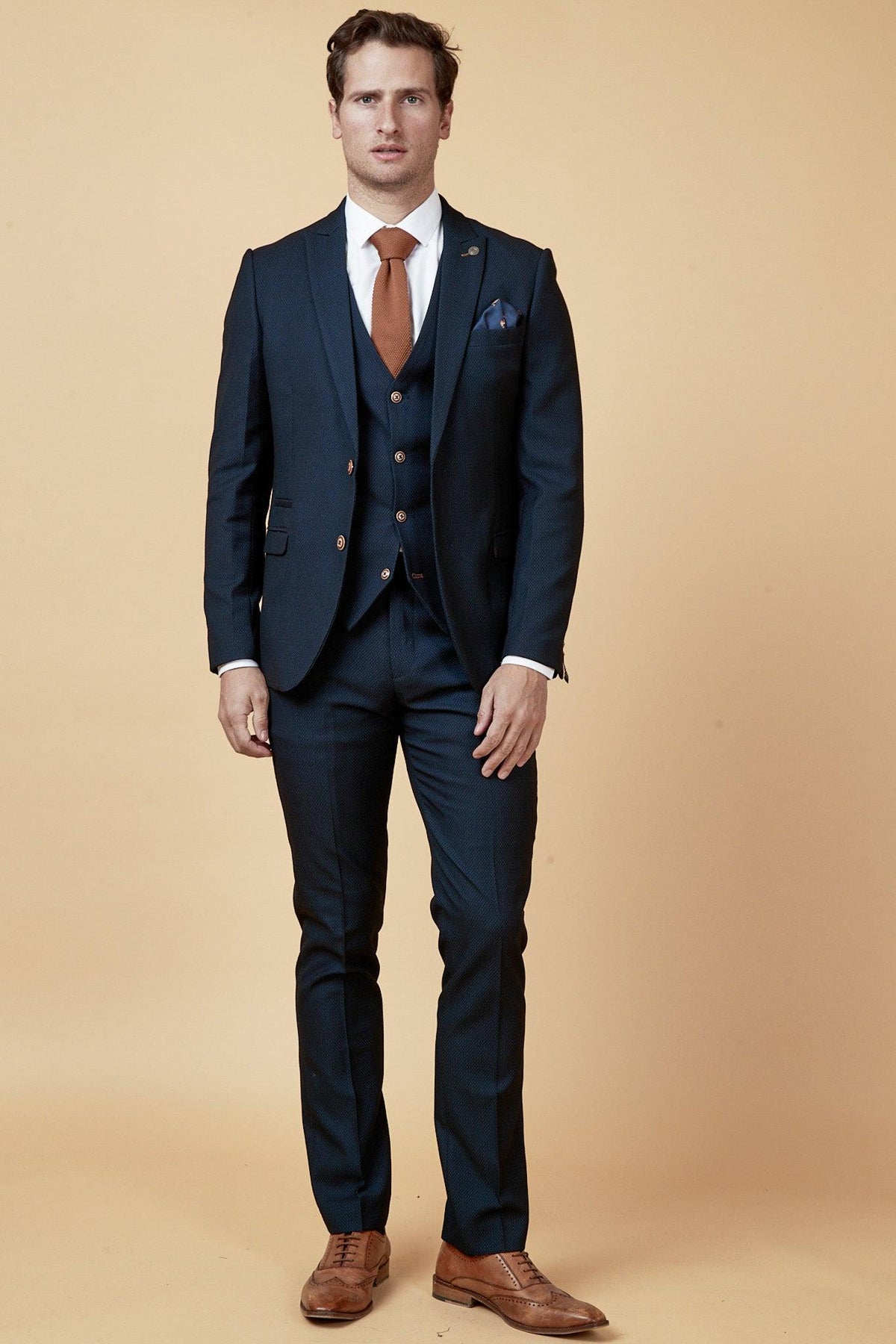A man in the marcdarcy Manchester United Treble Winner Denis Irwin Max Navy Suit stands against a tan background, featuring a white shirt, orange tie, brown shoes, and a camouflage print pocket square.