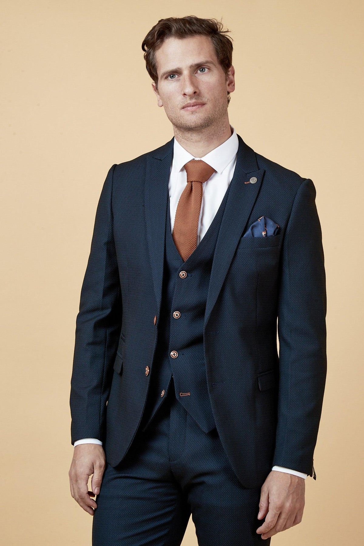 A man wears the marcdarcy Manchester United Treble Winner Denis Irwin in Max Navy Suit, styled with a white shirt, burnt orange knitted tie, and camouflage print pocket square against a beige background.