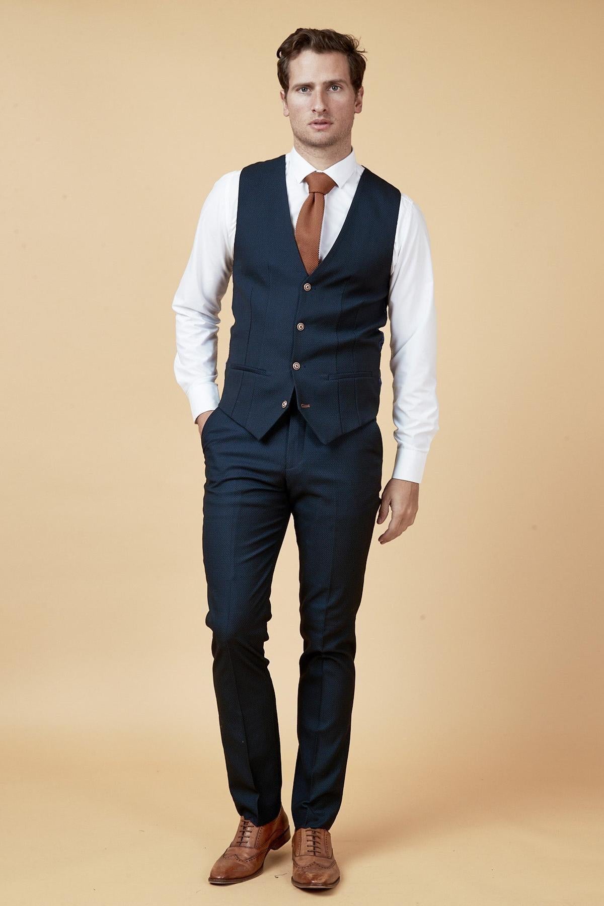 A man stands in a dark blue suit, white shirt, rust-colored tie, and the marcdarcy MAX Navy Single Breasted Waistcoat, with his hand in his pocket.