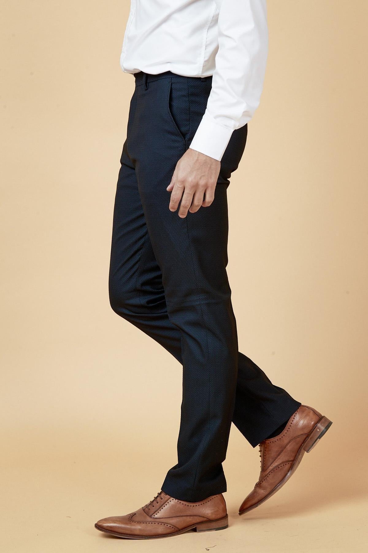 A man in a white dress shirt and marcdarcy's MAX Navy Blue Trousers with Contrast Buttons, paired with brown leather wingtip shoes, stands against a light tan background—a true formalwear staple.