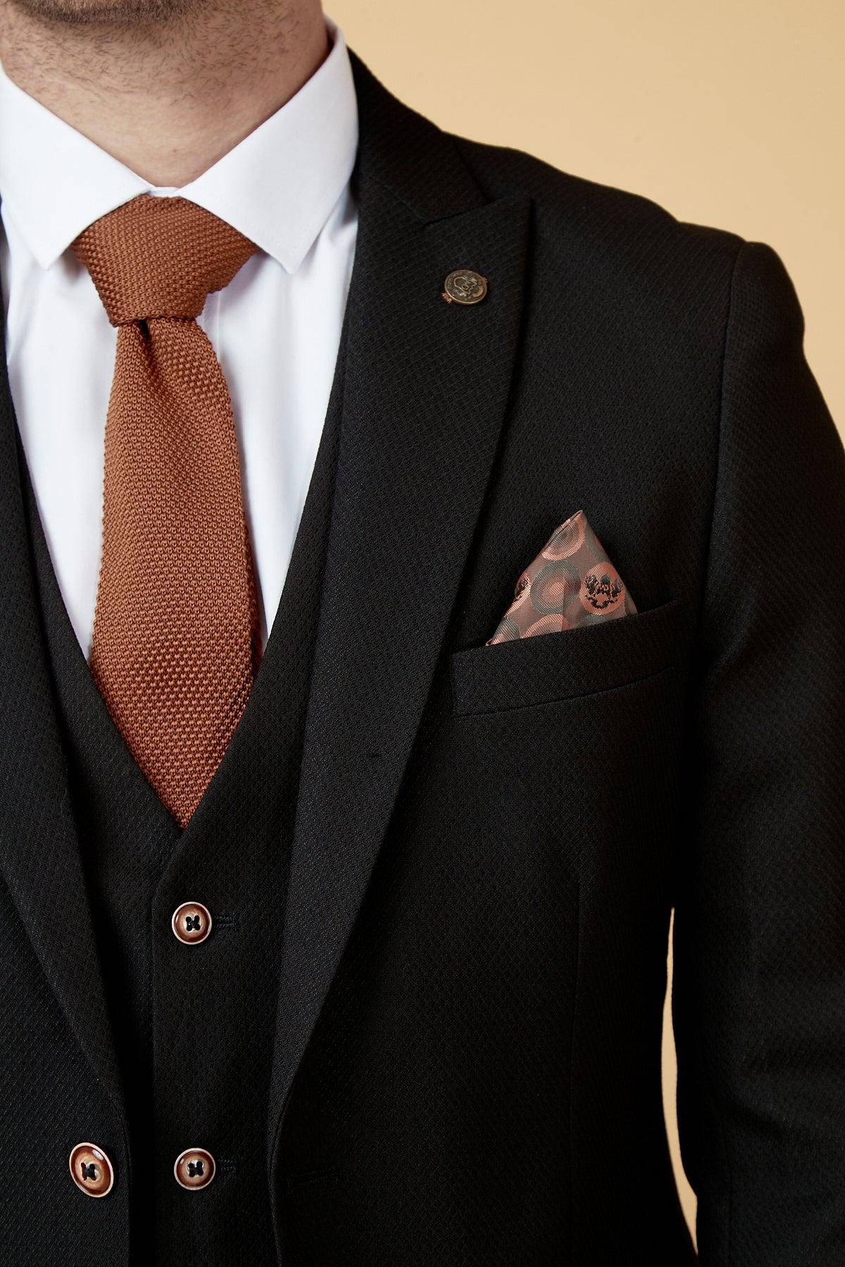 A man wears the marcdarcy MAX black three piece suit with contrast buttons, paired with a white shirt, textured brown tie, camouflage pocket square, and a lapel pin.
