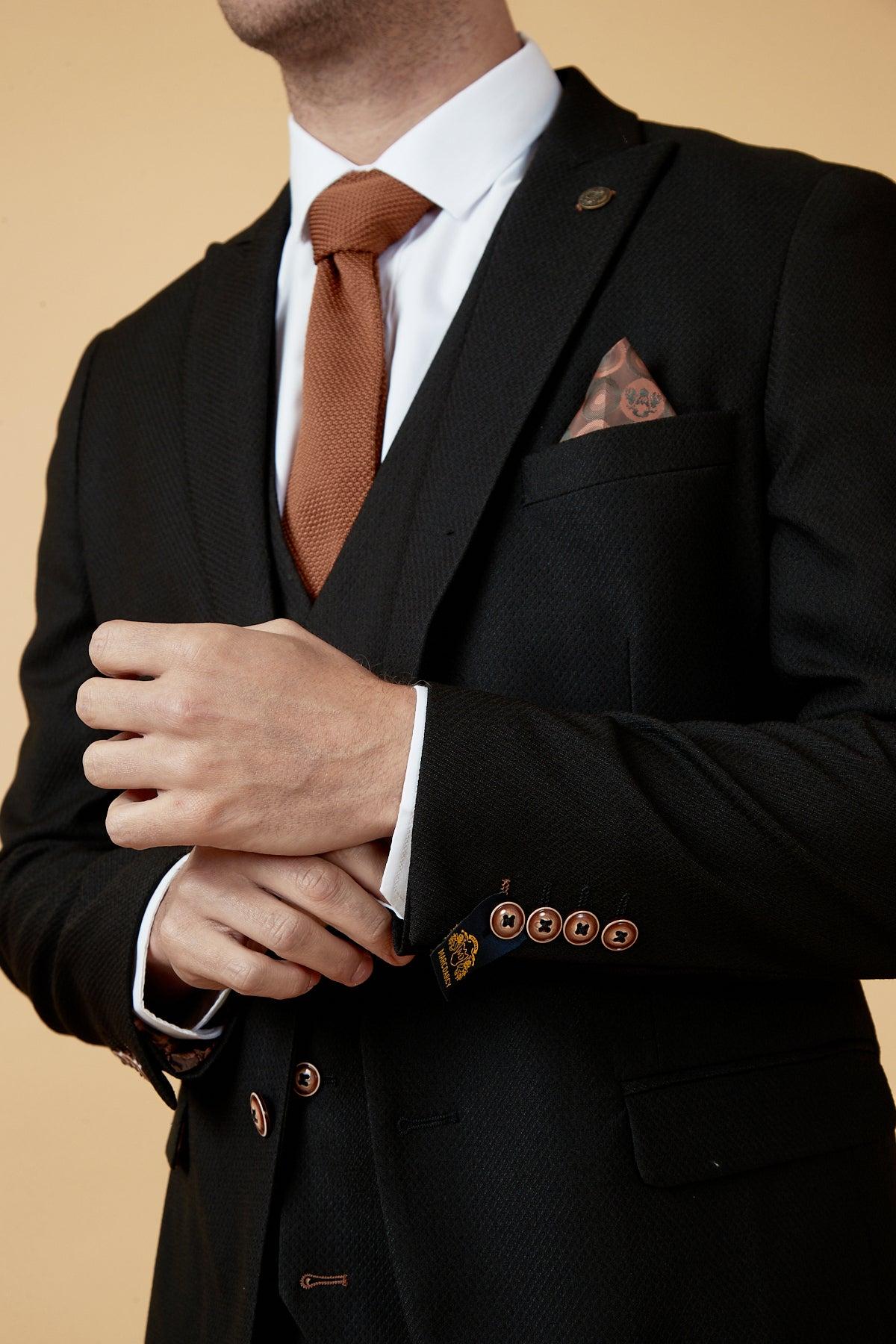 A man in the marcdarcy MAX Black Three Piece Suit with Contrast Buttons adjusts his cuff, revealing a camouflage pocket square tucked into his breast pocket above a white shirt and rust-colored tie.