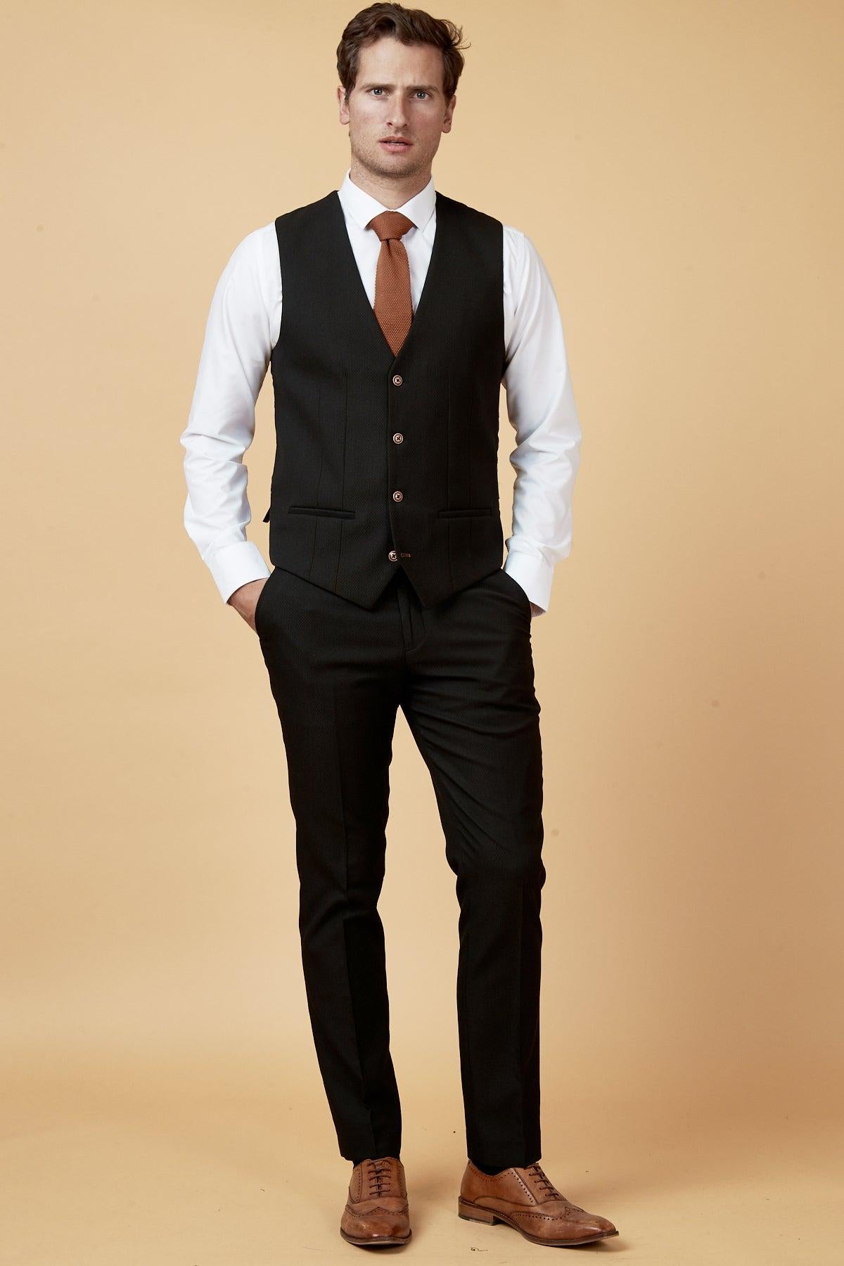 A man stands against a beige background in a slim fit white shirt, black pants, brown tie and dress shoes, styled with the marcdarcy MAX Black Single Breasted Waistcoat. His hands are in his pockets as he looks at the camera.