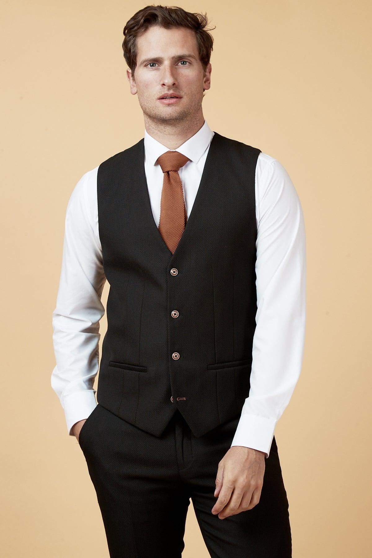A man wears the marcdarcy MAX Black Single Breasted Waistcoat over a white shirt with a rust tie, hand in pocket, looking forward against a tan background—a sharp upgrade for any workwear wardrobe.