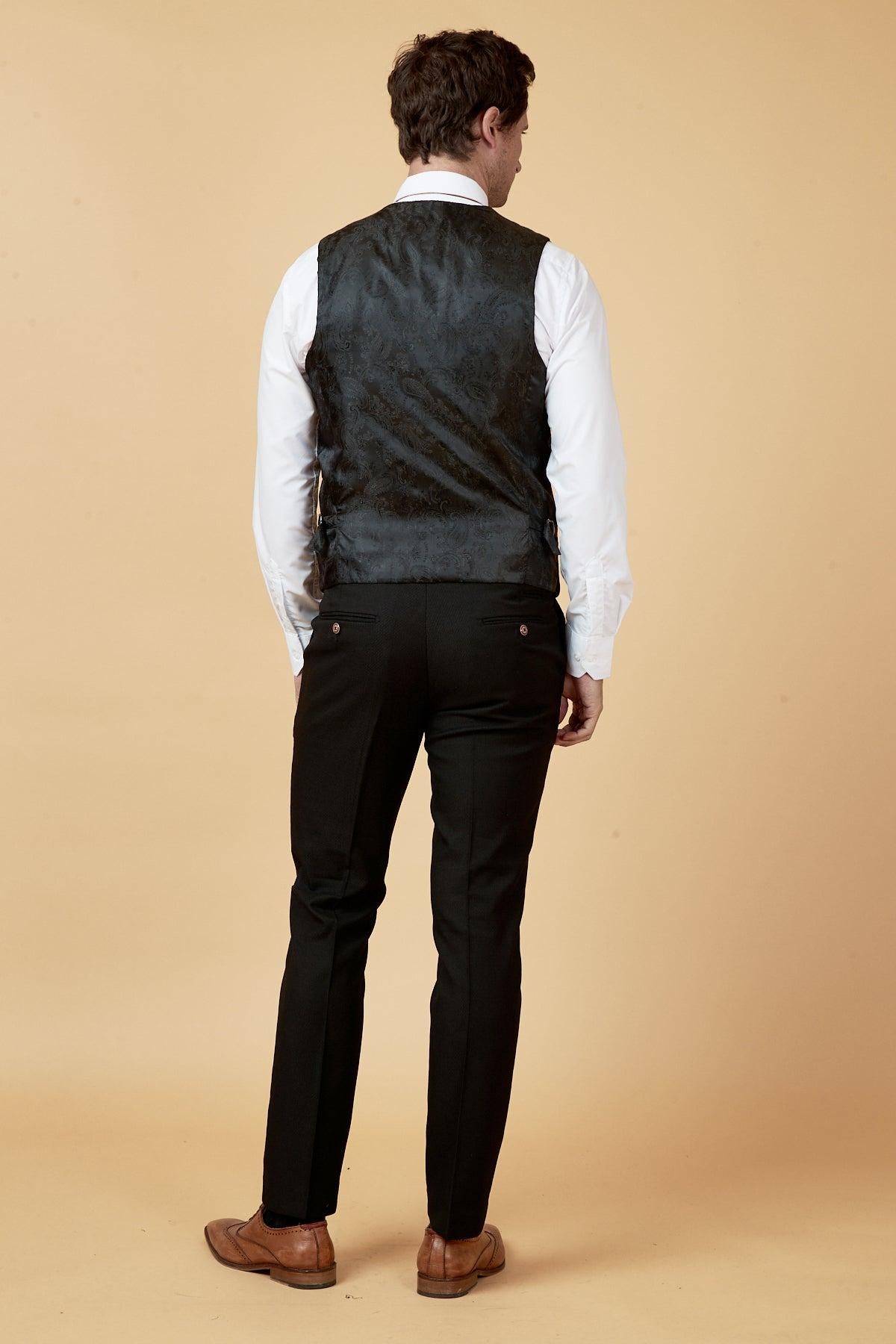A man stands facing away in black trousers, a white dress shirt, brown dress shoes, and the marcdarcy MAX Black Single Breasted Waistcoat with a tailored fit. The background is plain beige.