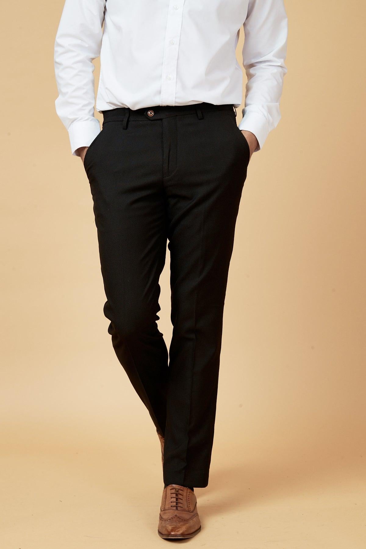 A model in a white shirt and marcdarcy’s MAX Black Trousers with Contrast Buttons stands legs crossed, hands in pockets, wearing brown dress shoes against a beige backdrop.