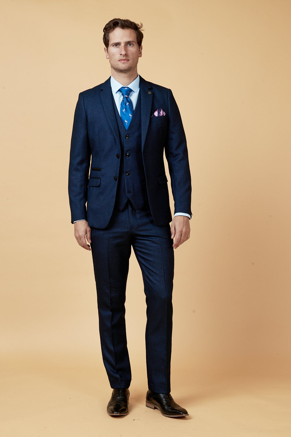 A man with short brown hair stands against a light beige background wearing the marcdarcy CALLUM Blue Three Piece Suit, styled with a white shirt, blue patterned tie, pocket square, and black dress shoes for a modern formal look.