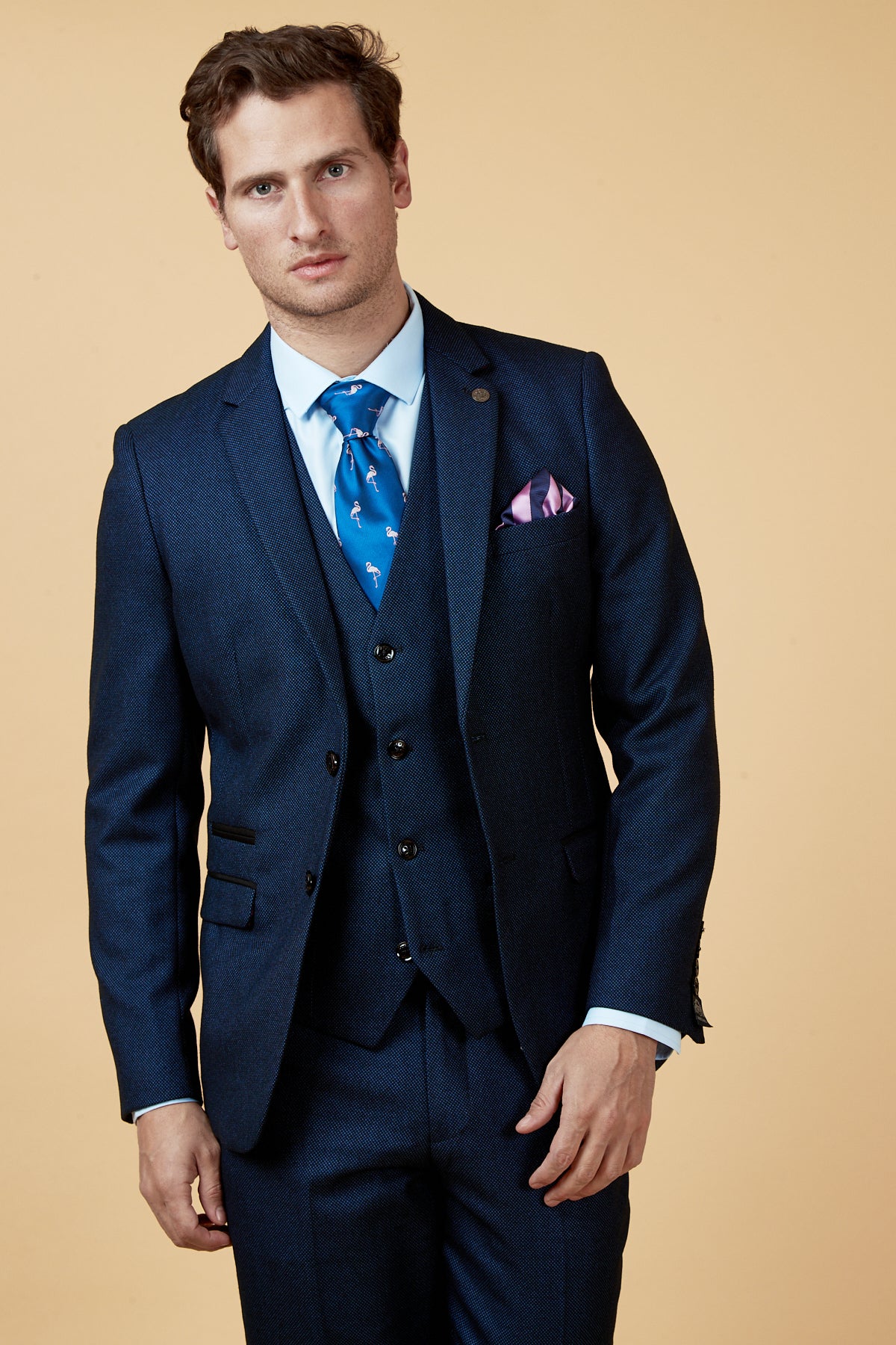 A man with short brown hair, wearing the marcdarcy CALLUM - Blue Blazer with a pink pocket square, light blue dress shirt, and blue patterned tie stands against a beige background.