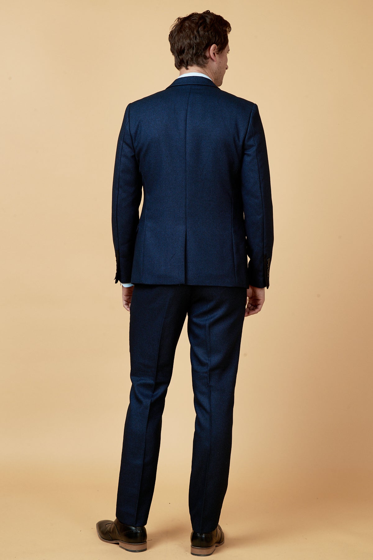 A man models the marcdarcy CALLUM Blue Three Piece Suit with a white shirt and brown shoes, standing with his back to the camera against a beige background, highlighting classic men’s formalwear.