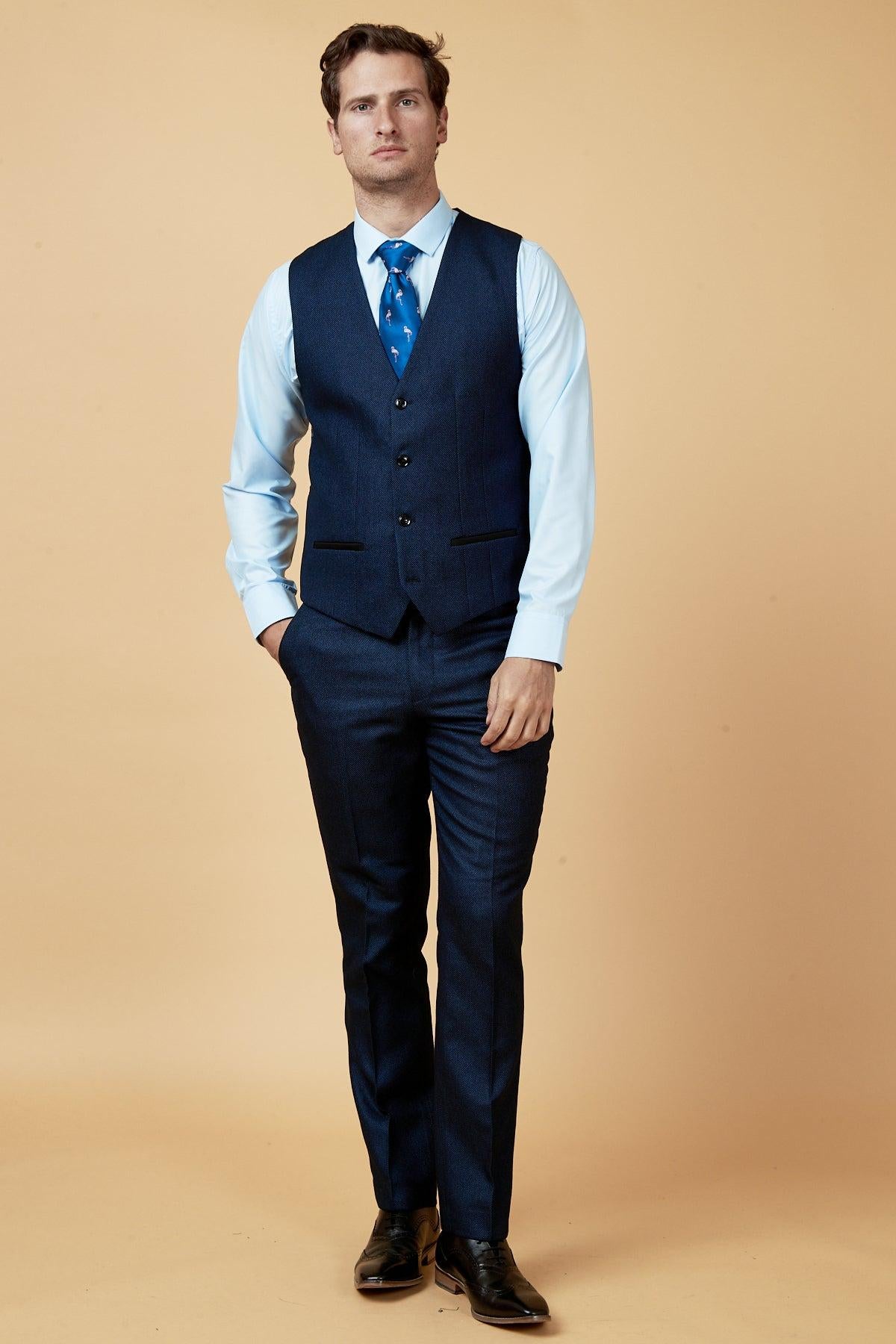 A man wears the marcdarcy CALLUM blue waistcoat with a light blue shirt and a blue flamingo-patterned tie, standing with one hand in his pocket against a peachy background—a sharp choice for any work wardrobe.