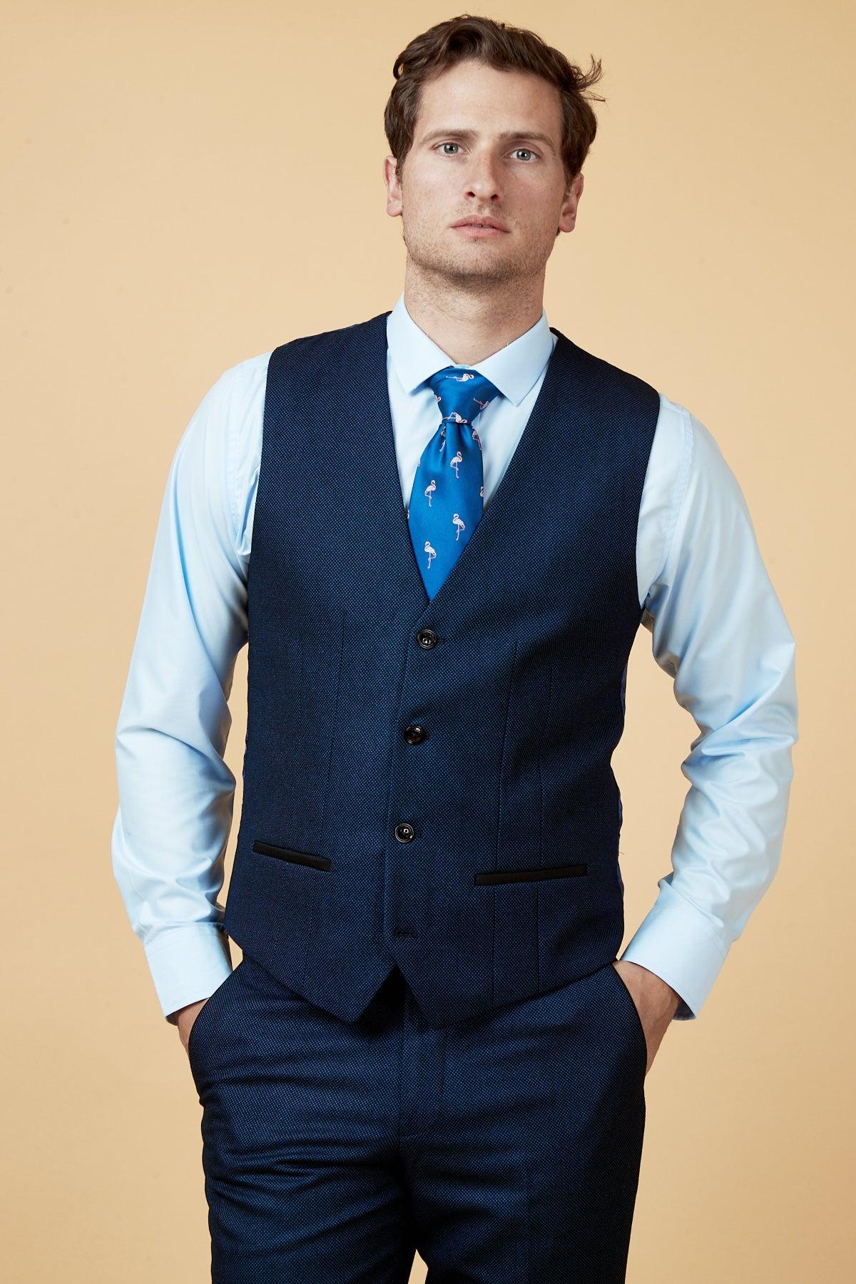 A man wears the marcdarcy CALLUM Blue Waistcoat with matching tailored fit pants, a light blue dress shirt, and a blue patterned tie, standing hands in pockets against a plain beige background.