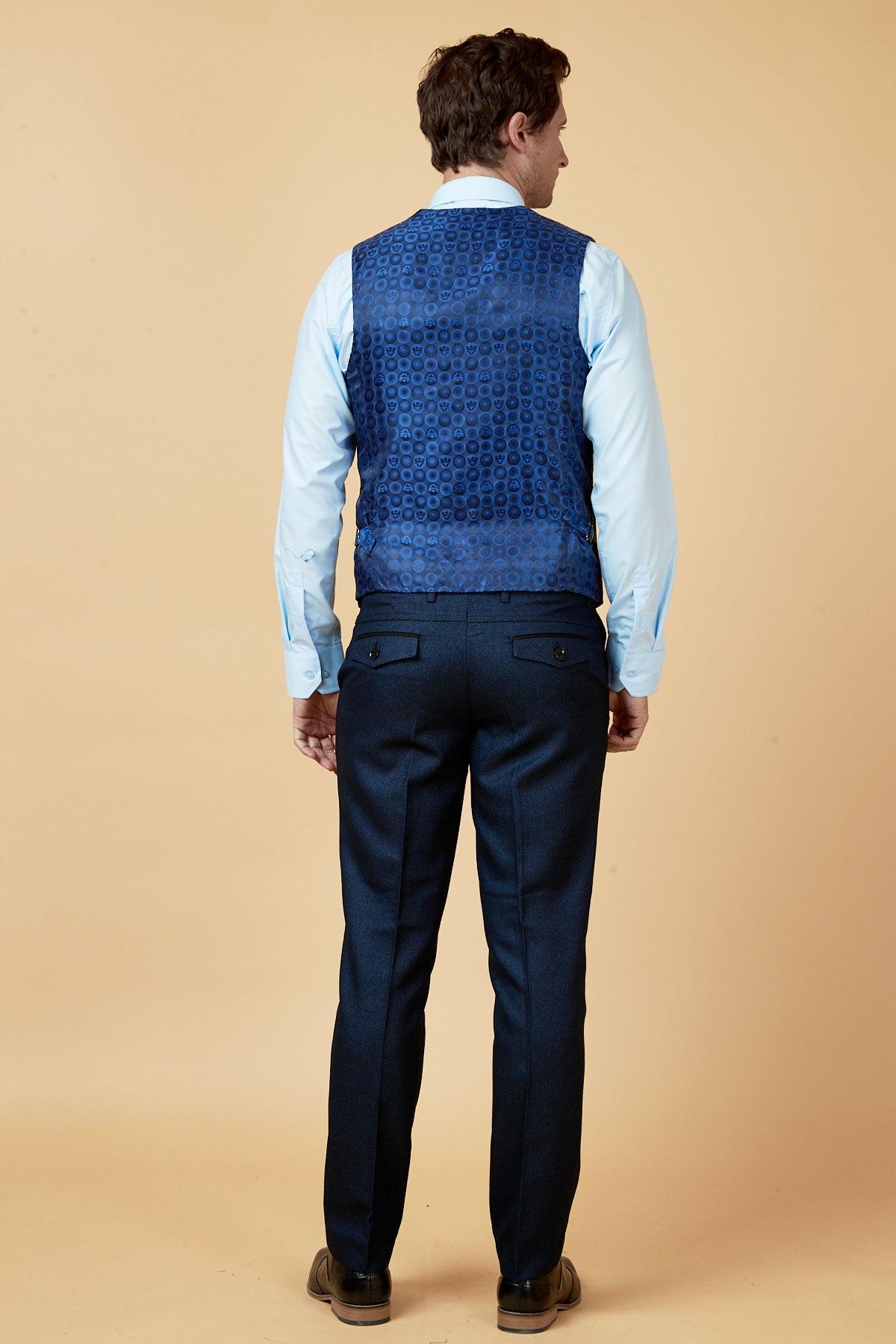 A man faces away from the camera, dressed in dark pants, brown shoes, a light blue shirt, and the marcdarcy CALLUM - Blue Waistcoat against a beige background—a sharp look for any professional setting.