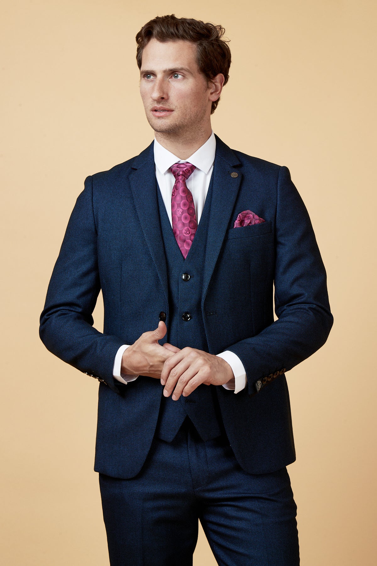 A man wears the marcdarcy CALLUM - Blue Blazer three-piece suit with a white shirt, pink patterned tie, and matching pocket square against a beige background—an ideal choice for refined formal events.