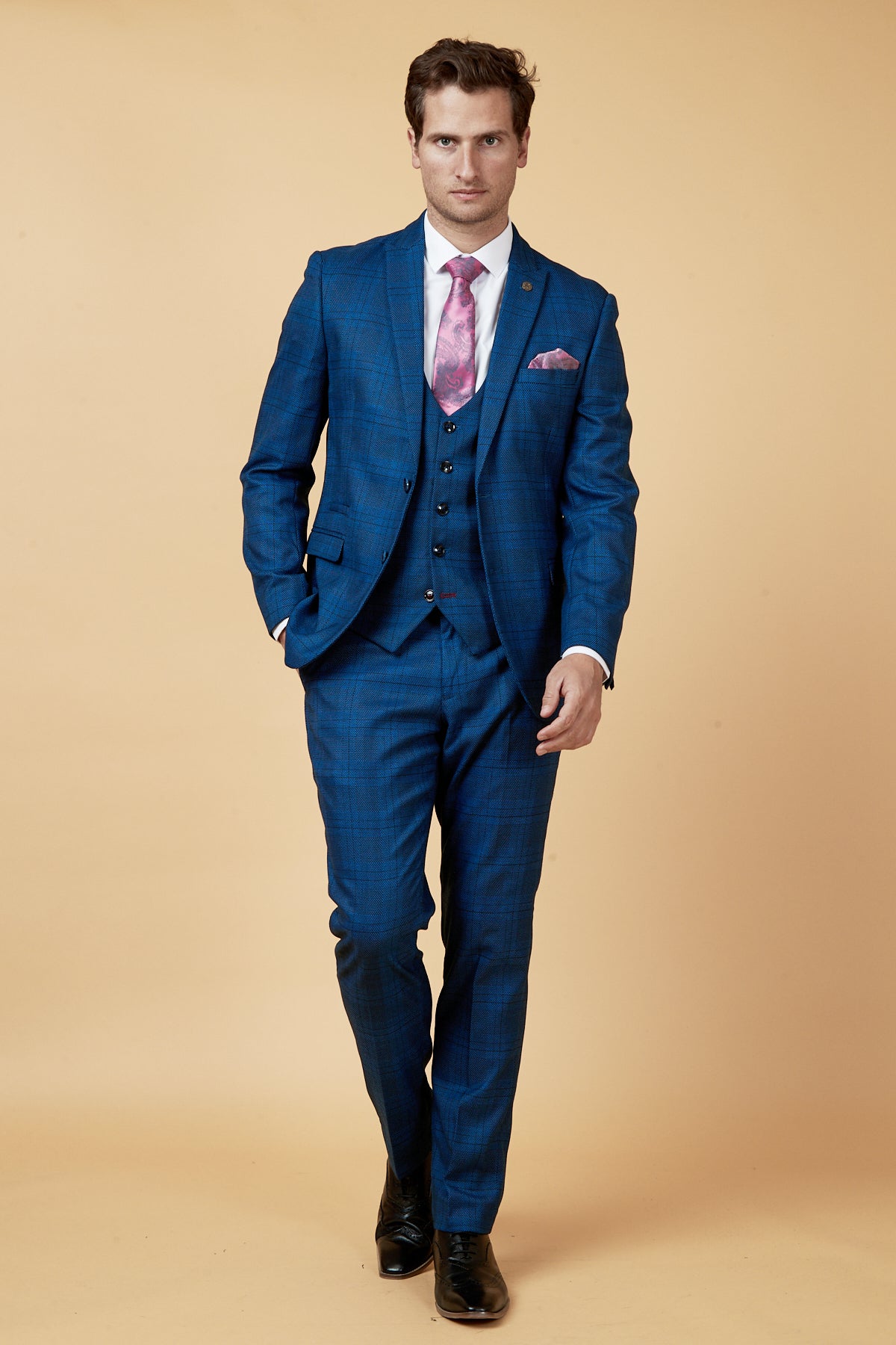 A man in the marcdarcy JERRY Blue Check Suit with single breasted waistcoat, styled with a pink patterned tie and pocket square, walks confidently against a beige background, highlighting its sharp slim fit.